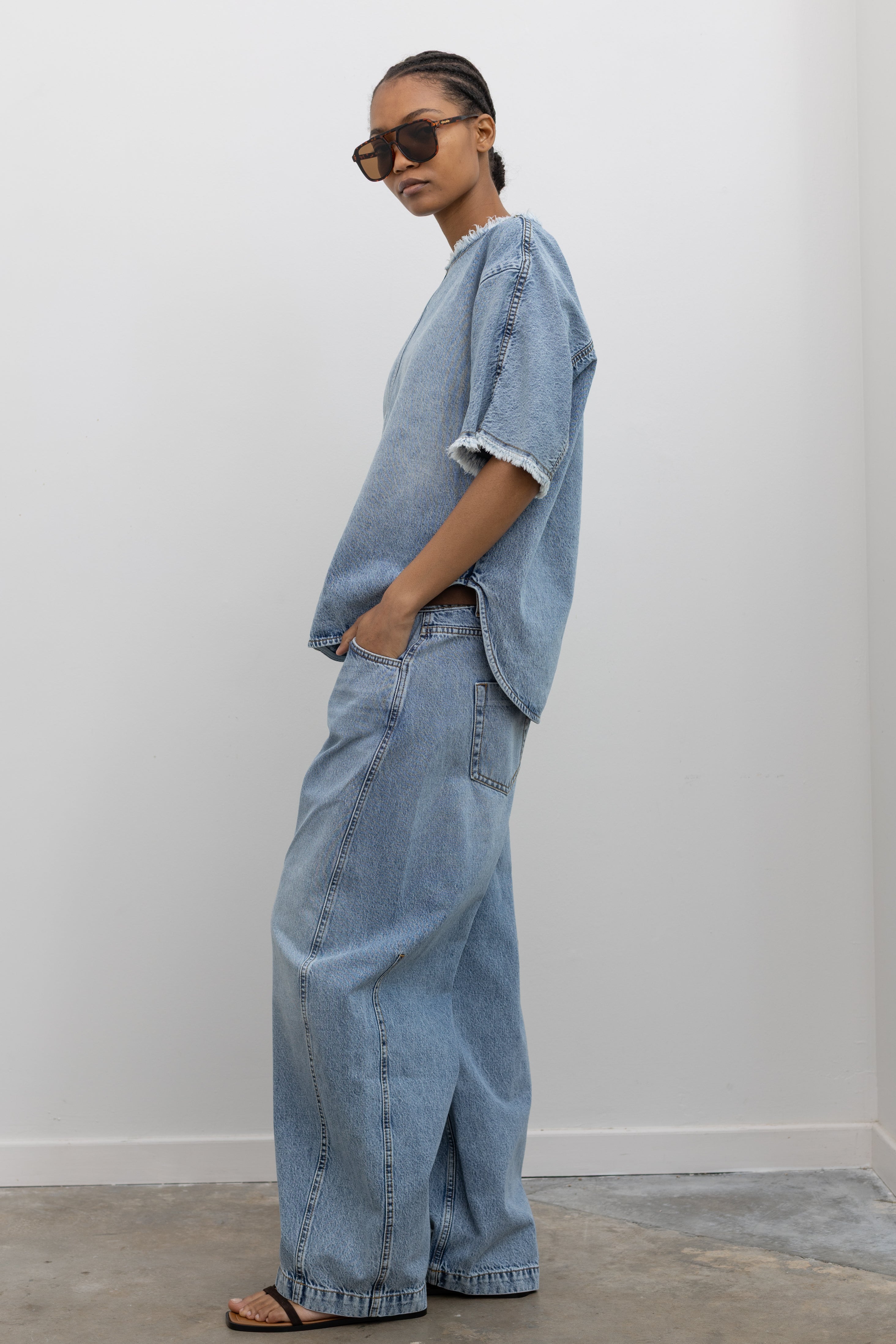 Full length side view of a woman posing wearing elevated casual spring street style fashion in a Tela Light Blue Denim Oversized Short Sleeve Shirt featuring a clean light-blue wash, distinct raw-edge detailing, and a minimalist back neck closure paired with Tela light-blue wash baggy denim jeans and strappy sandals at an Alas Eius store. 