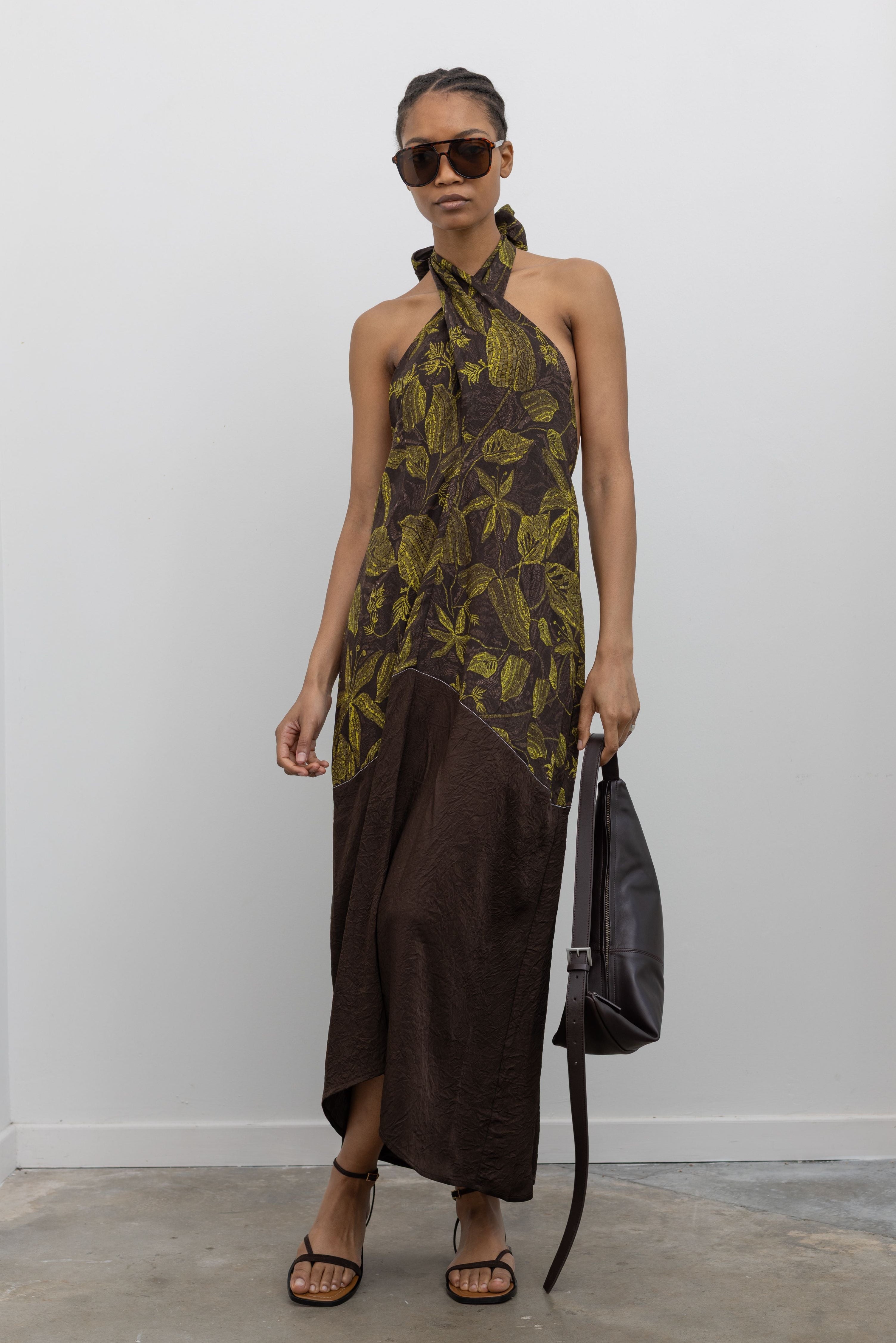Full length front view of a woman wearing the perfect summer vacation dress in a Tela label olive green botanical print halter neck midi dress with a contrasting chocolate brown hem paired with ATP strappy leather sandals and a leather St. Agni bag at an Alas Eius Store.
