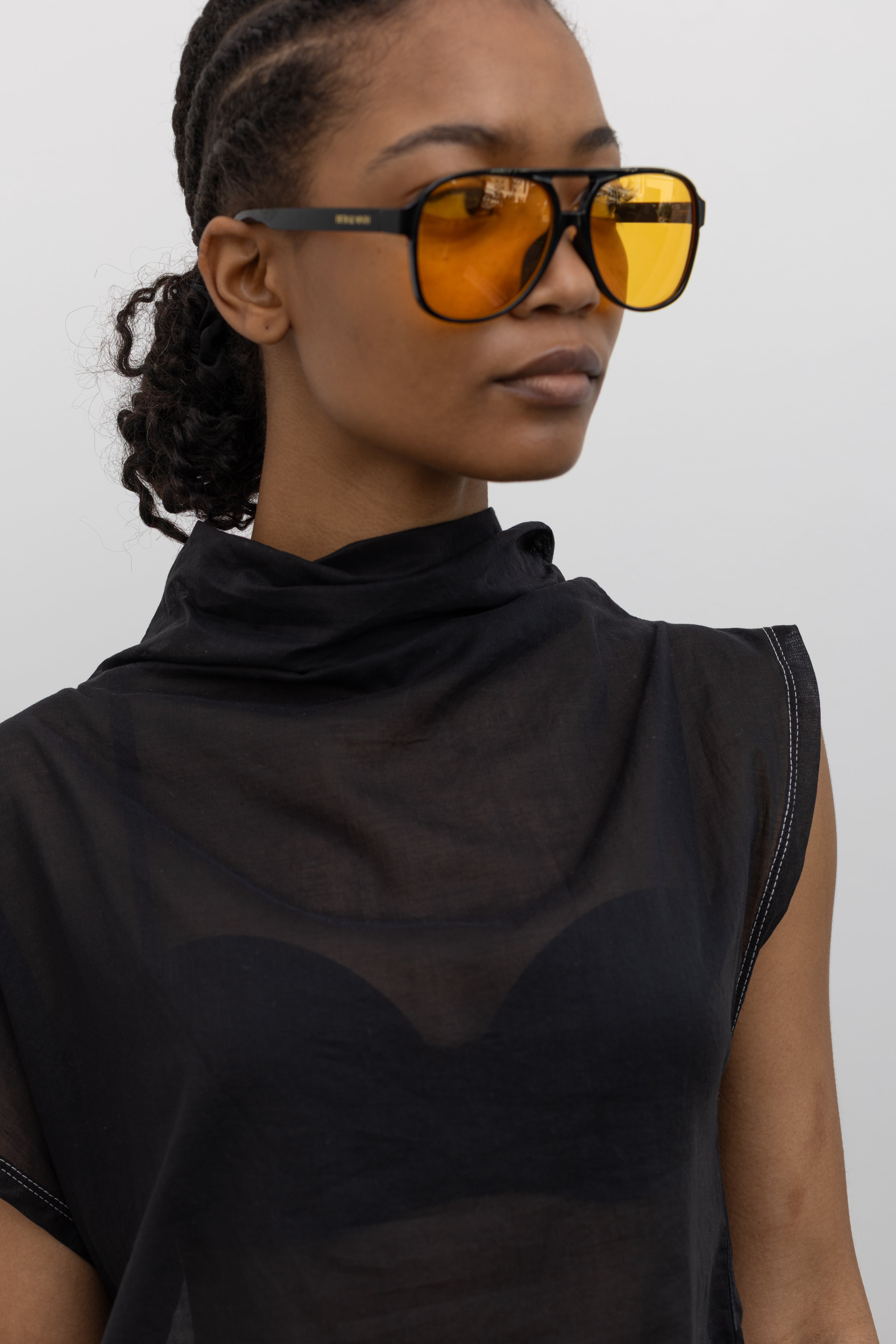 Close-up front view of a woman posing wearing the perfect date night top in a Tela brand semi-sheer lightweight washed muslin black top featuring a soft draped asymmetrical neckline paired with a classic wide black leather belt on black wide leg trousers at an Alas Eius store. 