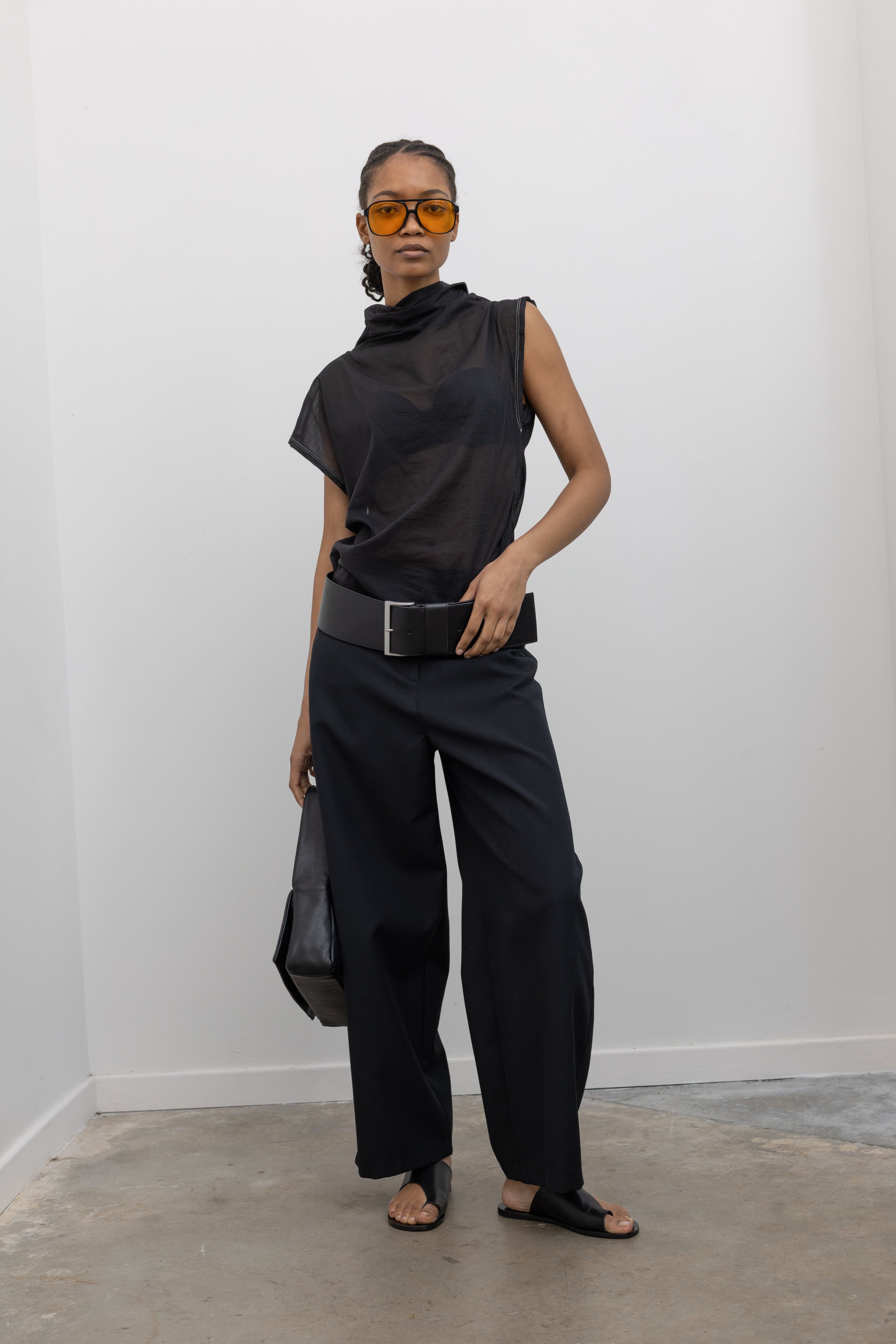Full length front view of a woman posing wearing a Tela brand semi-sheer lightweight washed muslin black top featuring a soft draped asymmetrical neckline paired with a classic wide black leather belt on black wide leg trousers at an Alas Eius store. 