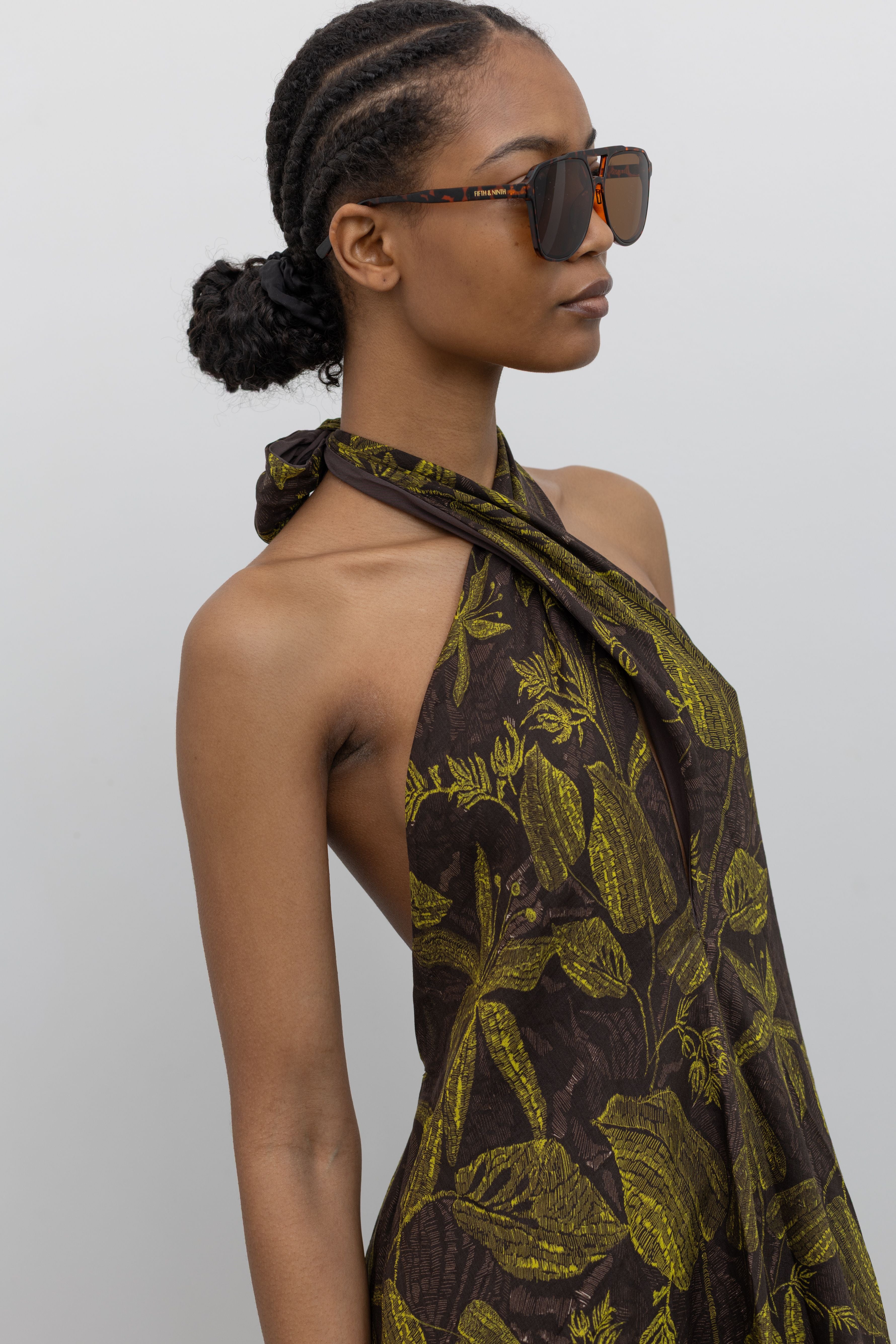 Close-up 3/4 view of a woman wearing the perfect summer vacation dress in a Tela label olive green botanical print halter neck midi dress with a contrasting chocolate brown hem at an Alas Eius Store.