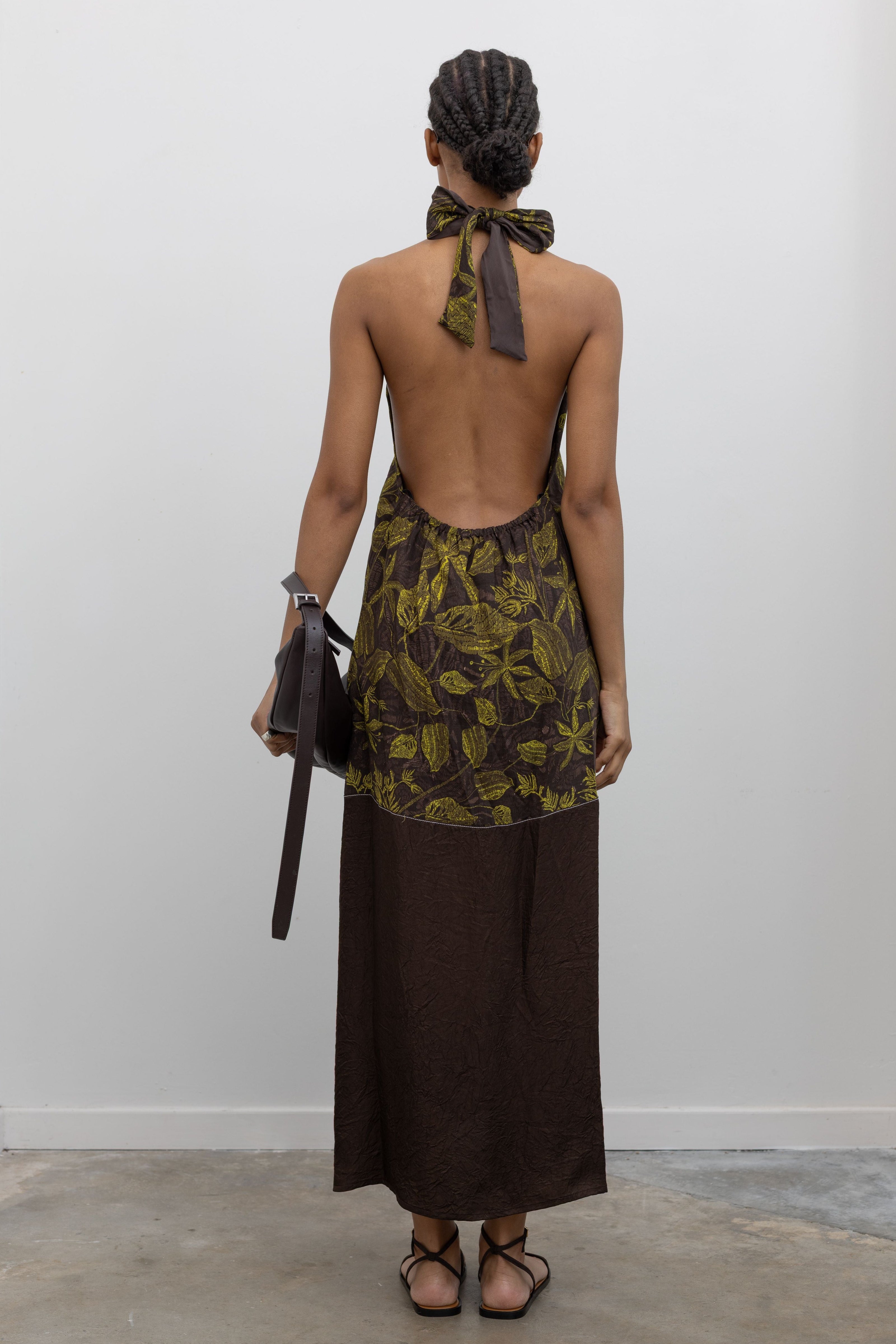 Full length back view of a woman standing wearing a summer essential  in a Tela label olive green floral print halter neck backless midi dress with a contrasting chocolate brown hem and paired with a St. Agni leather bag and strappy sandals at an Alas Eius Store.