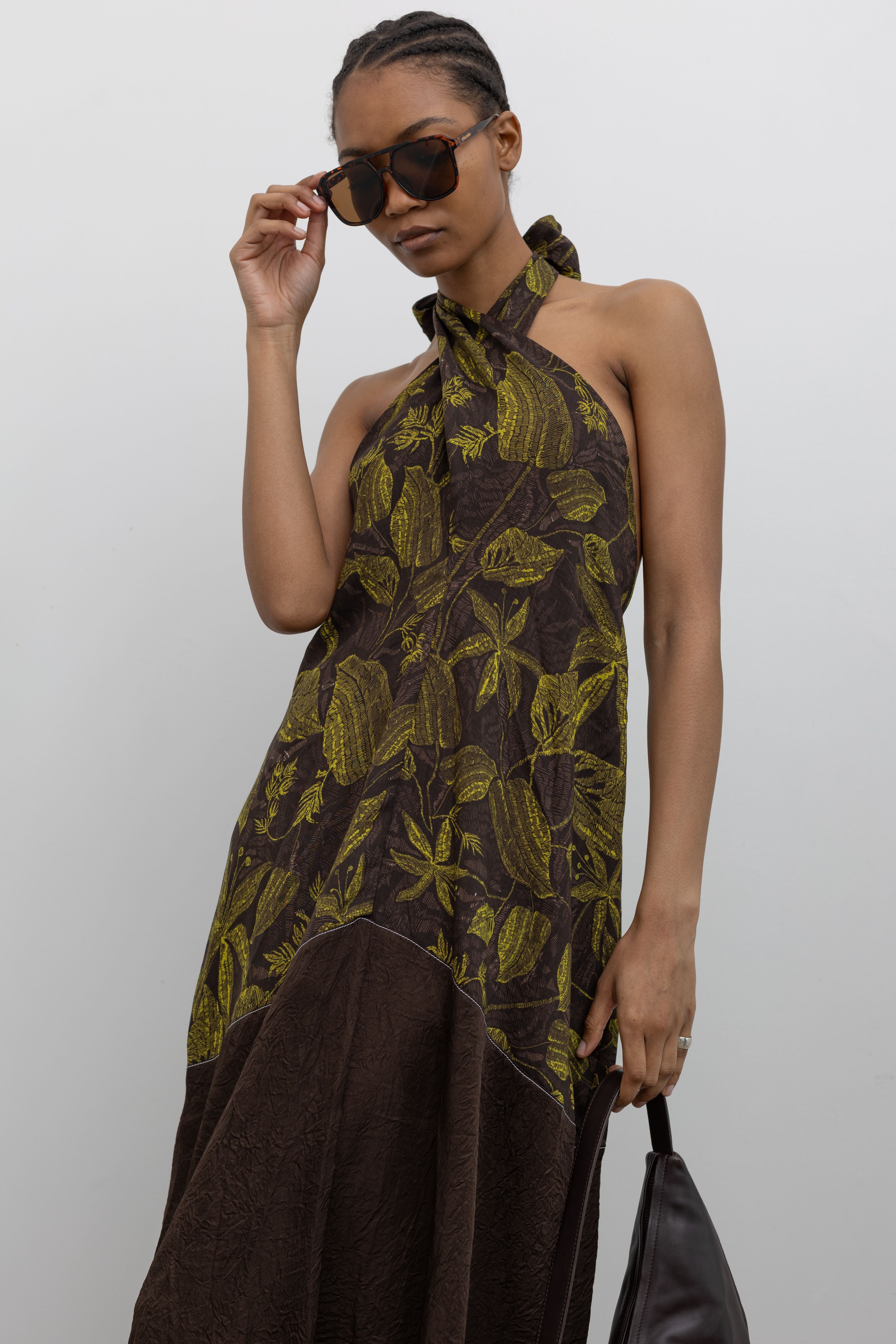 Woman posing with sunglasses wearing spring and summer fashion in a chic Tela label olive green floral print halter neck midi dress with a contrasting chocolate brown hem at an Alas Eius Store. 