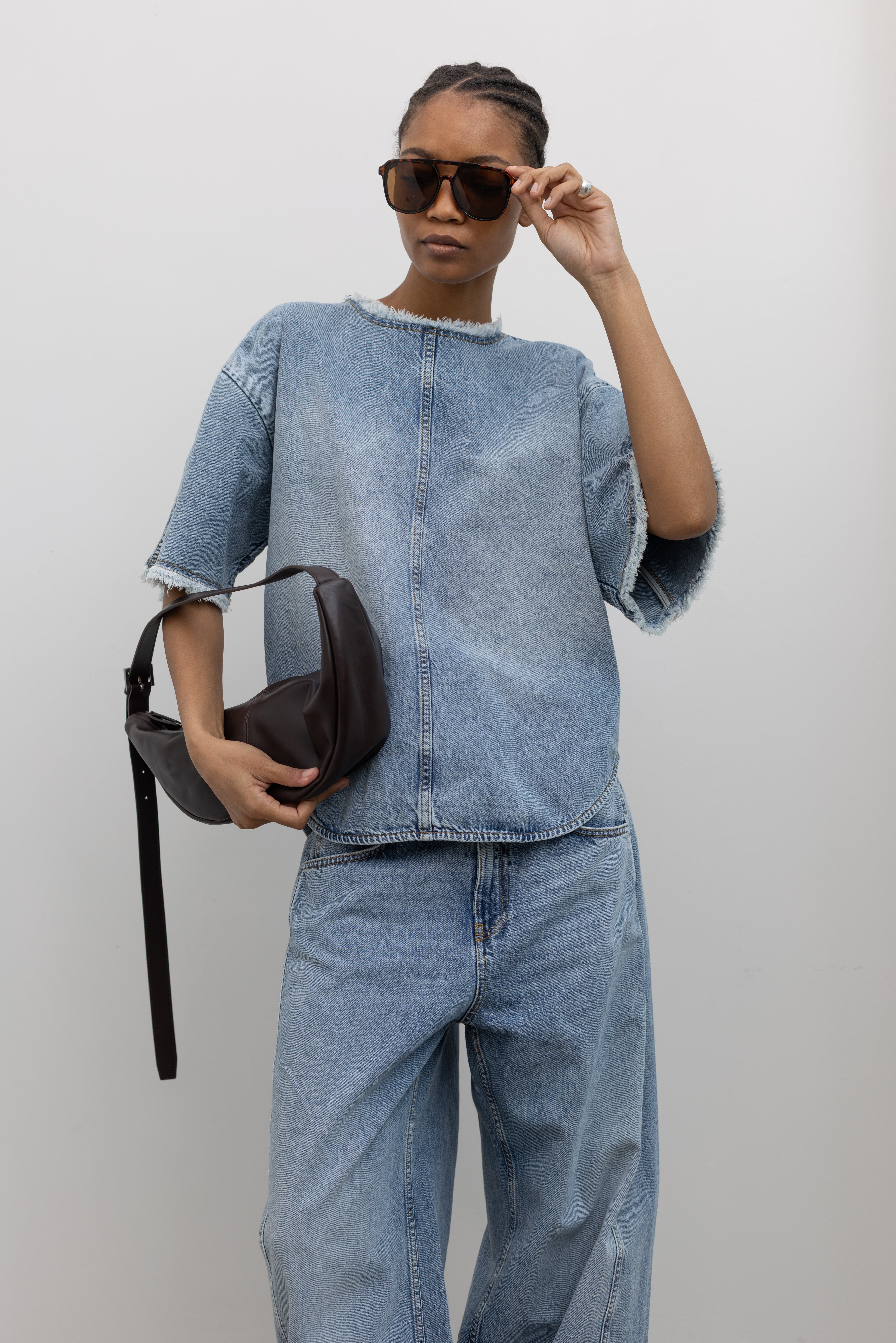 Woman posing with sunglasses and a St. Agni leather bag wearing an elevated and chic spring denim top by Tela featuring an oversized short-sleeve, a clean light-blue wash, distinct raw-edge detailing, and a minimalist back neck closure paired with light-blue wash denim jeans at Alas Eius. 