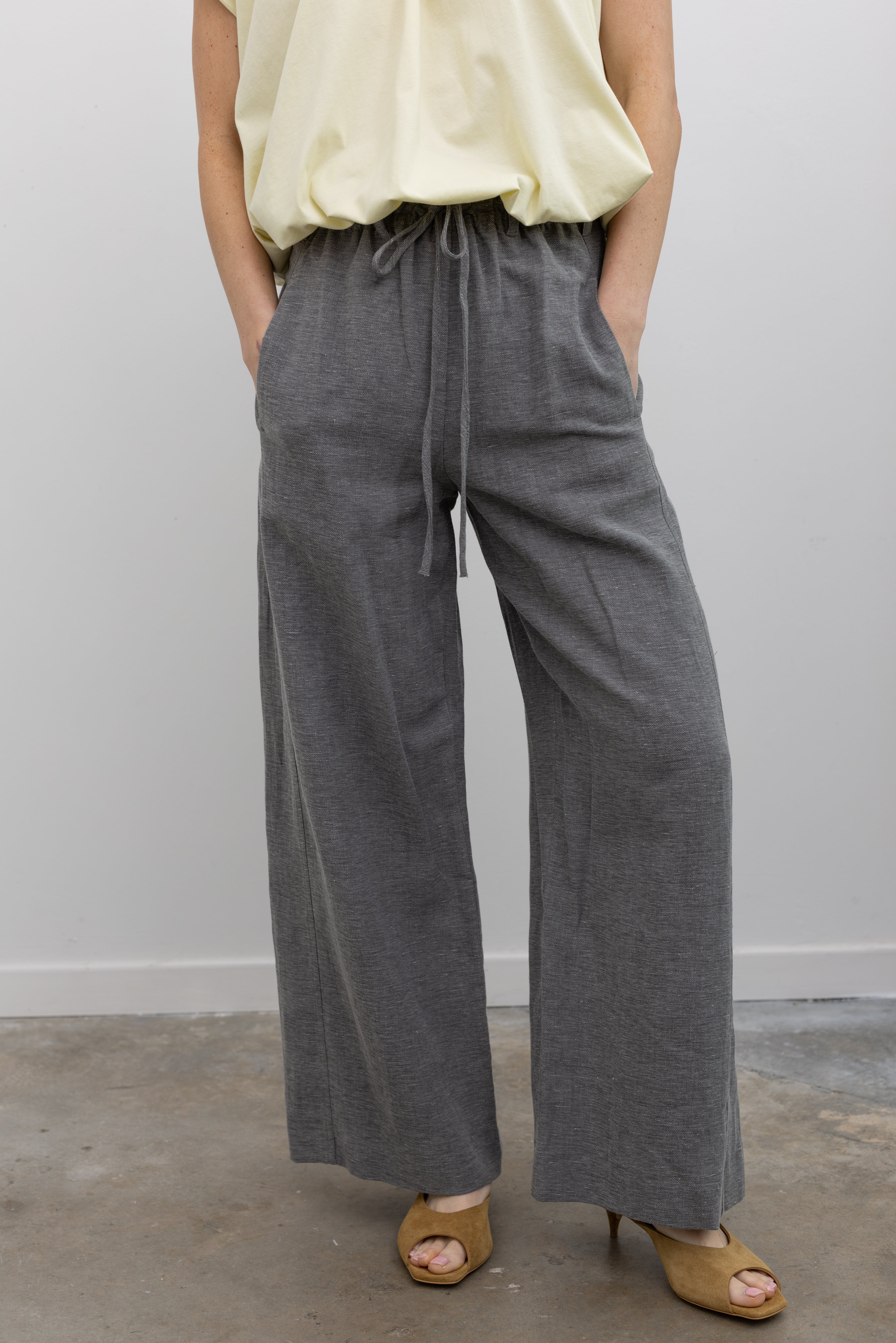 Woman standing wearing modern minimalist spring fashion in the Tela Lucio light gray drawstring pants featuring a wide leg and side seam pockets at an Alas Eius store. 