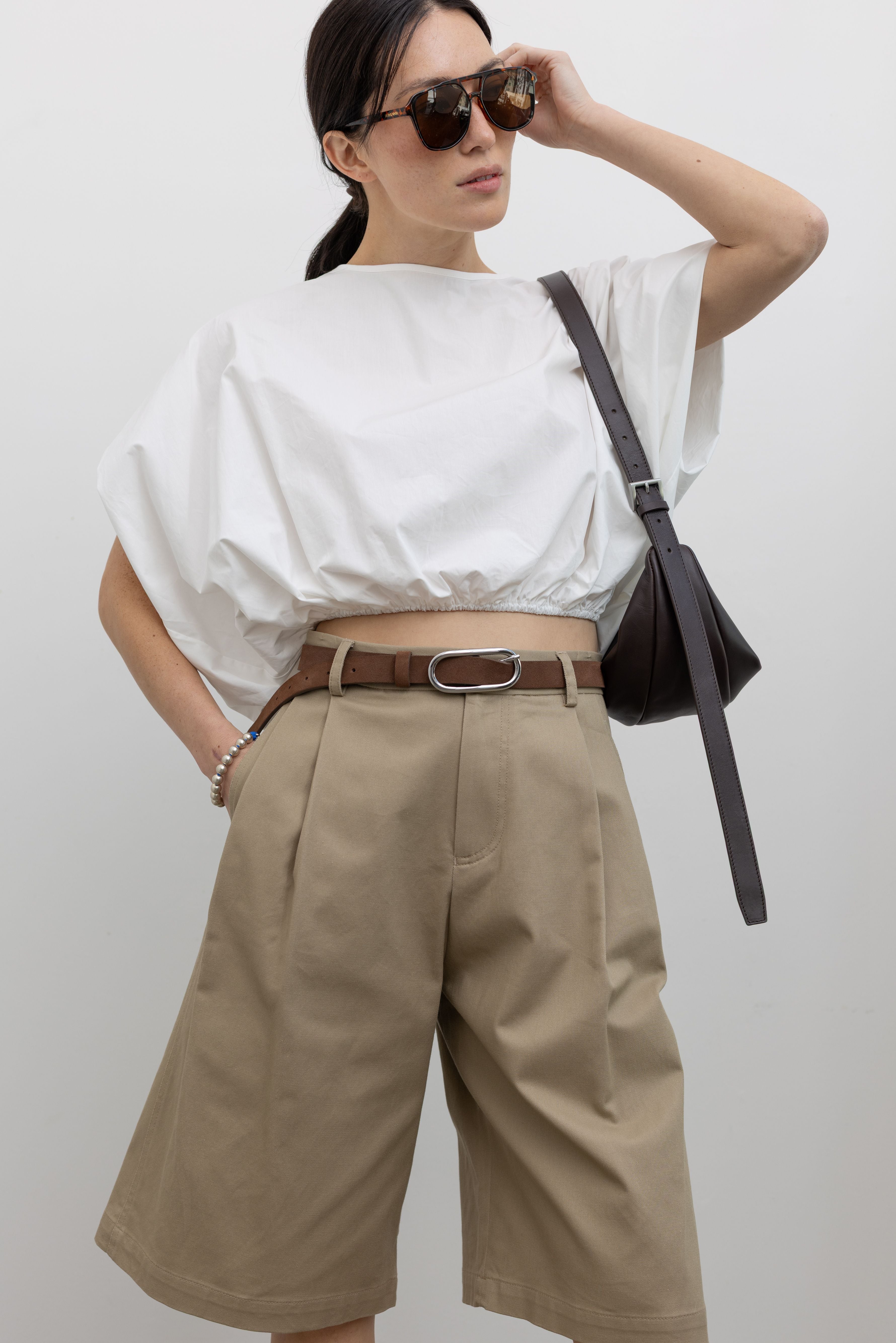 Close-up front view of a woman posing wearing a classic yet modern spring fashion in the Tela sand cotton gabardine Bermuda long short featuring a front pleat and side seam pockets paired with a classic brown leather belt, a crisp white top, a St. Agni leather bag, and white leather pointed toe kitten heels at an Alas Eius store. 