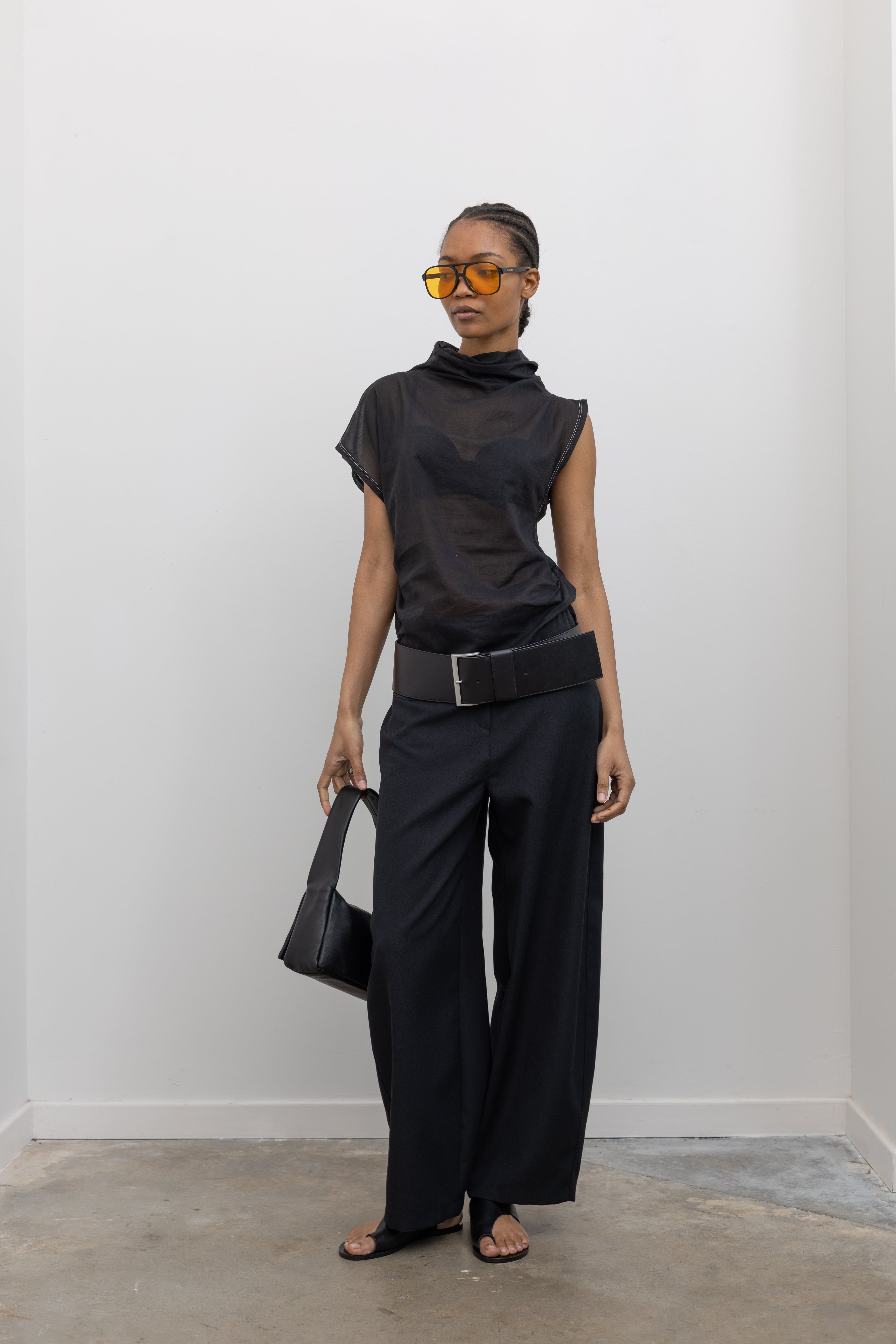 Front view of a woman posing wearing a chic and modern Tela brand semi-sheer lightweight washed muslin black top featuring a soft draped asymmetrical neckline paired with a classic wide black leather belt on black wide leg trousers at an Alas Eius store. 