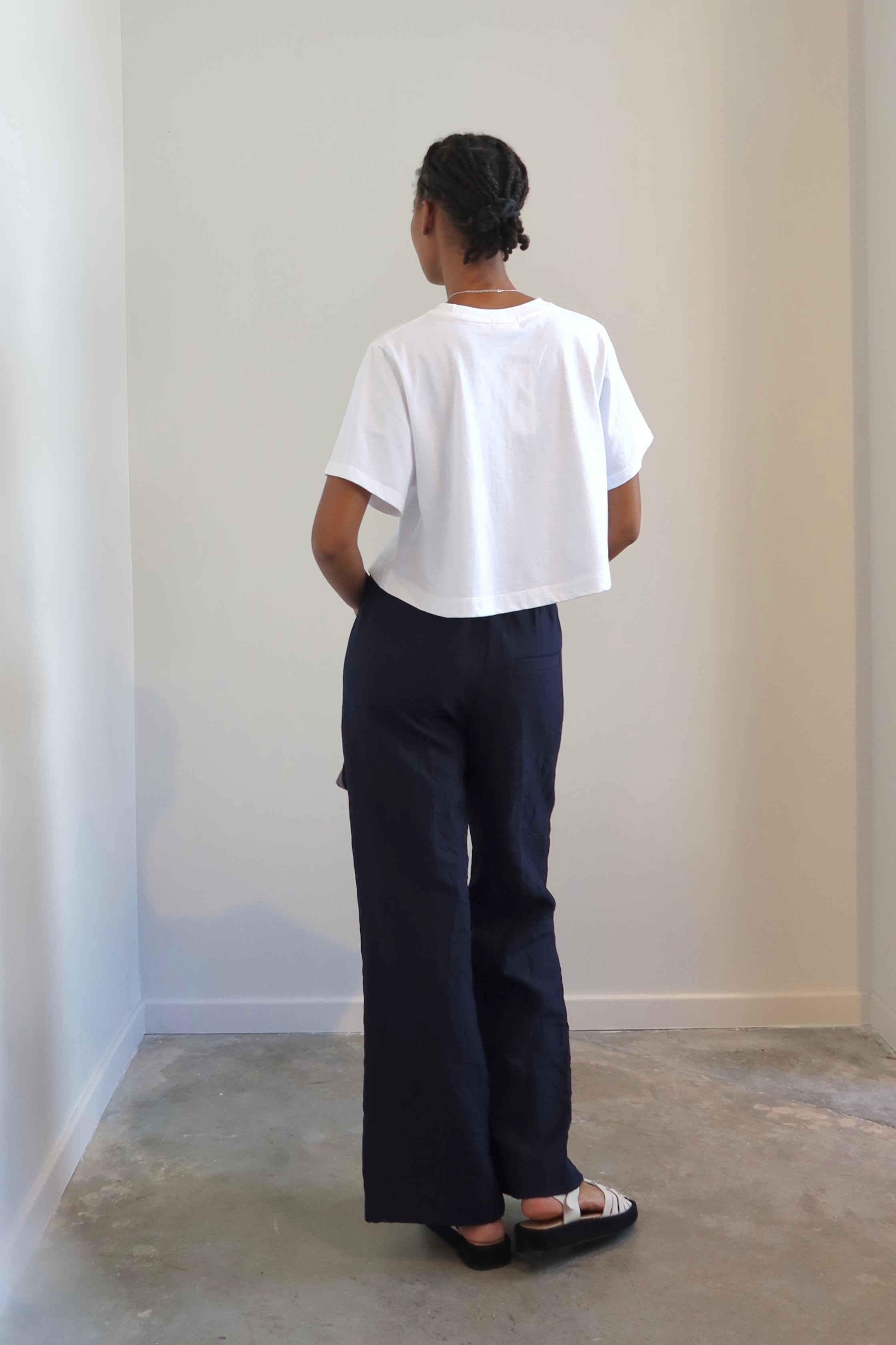 Back view of a Woman standing wearing Tela Radio Midnight Tailored Trousers featuring  an intentionally crinkled texture, elastic waistband, hidden drawstring, Mid-rise waist, and a slim fitting leg at Alas Eius.