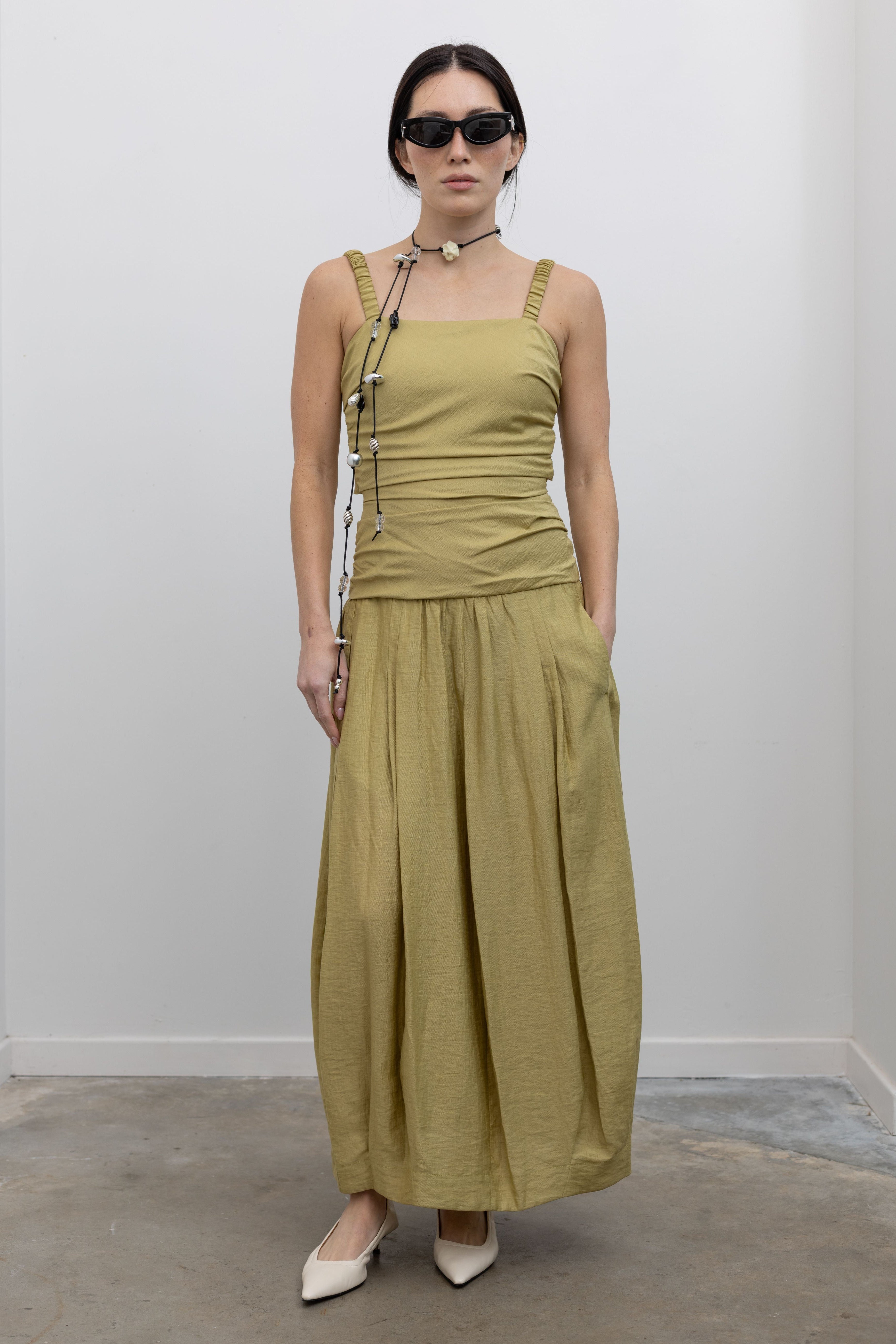 Full length front view of a woman standing wearing modern and elegant spring fashion in a Tela label linen blend pistachio green maxi dress with flattering ruching, delicate narrow shoulder straps, and a striking back cut-out detail with a bow closure paired with white leather slingback kitten heels at an Alas Eius store. 