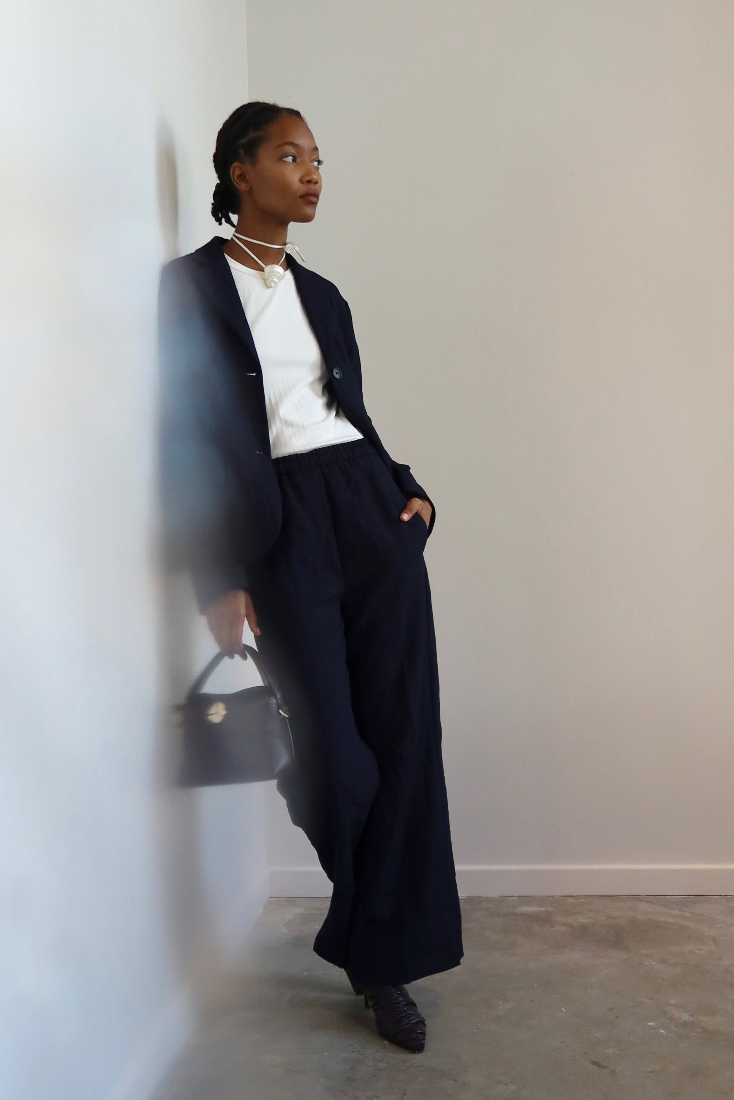Woman leaning against a wall wearing Tela Radio Midnight Tailored Trousers featuring an intentionally crinkled texture, elastic waistband, hidden drawstring, Mid-rise waist, and a slim fitting leg at Alas Eius.