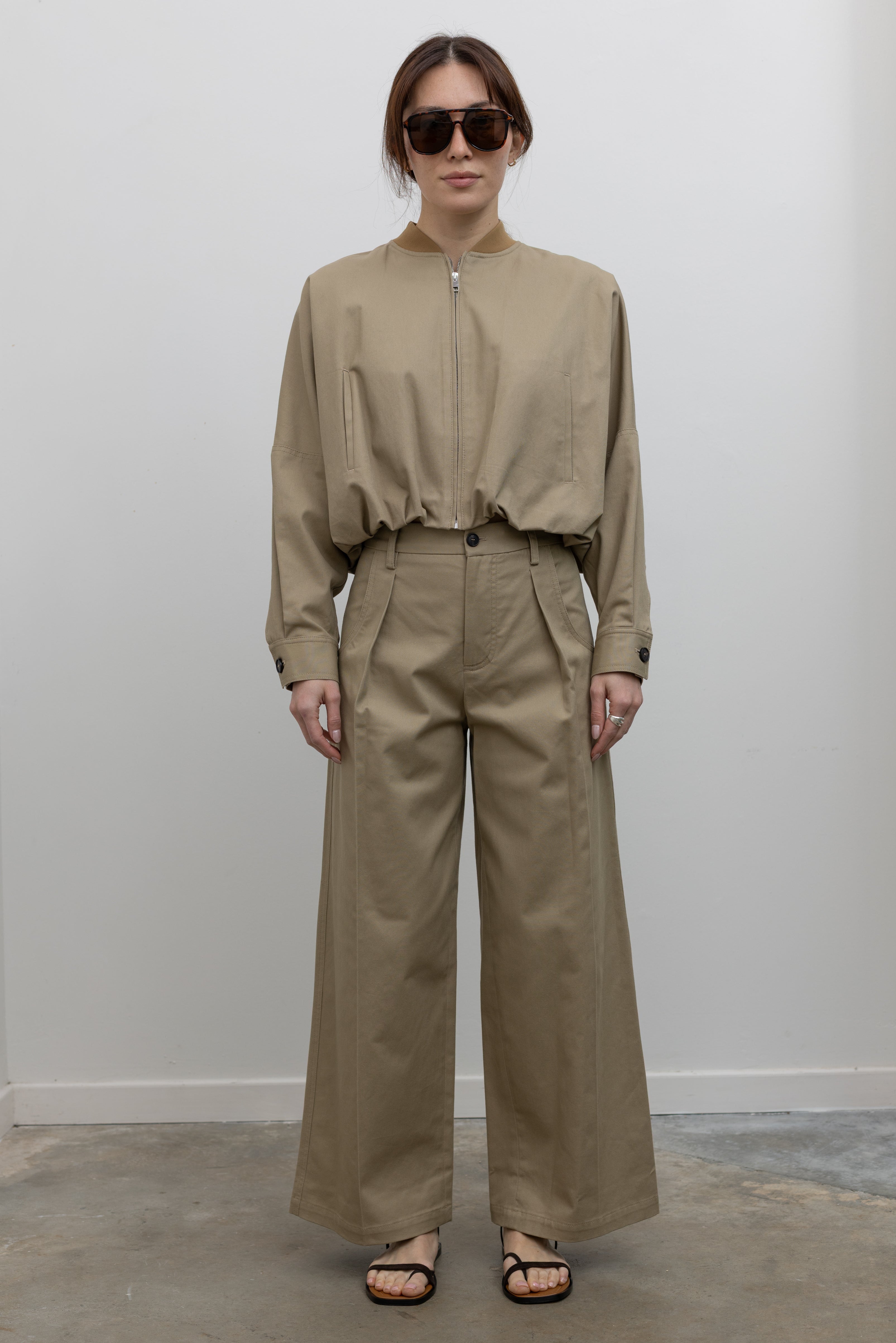 Woman standing wearing Spring fashion in a pair of Tela Rubicon Sand Cotton Gabardine Trousers and cropped jacket with sunglasses and sandals at Alas Eius