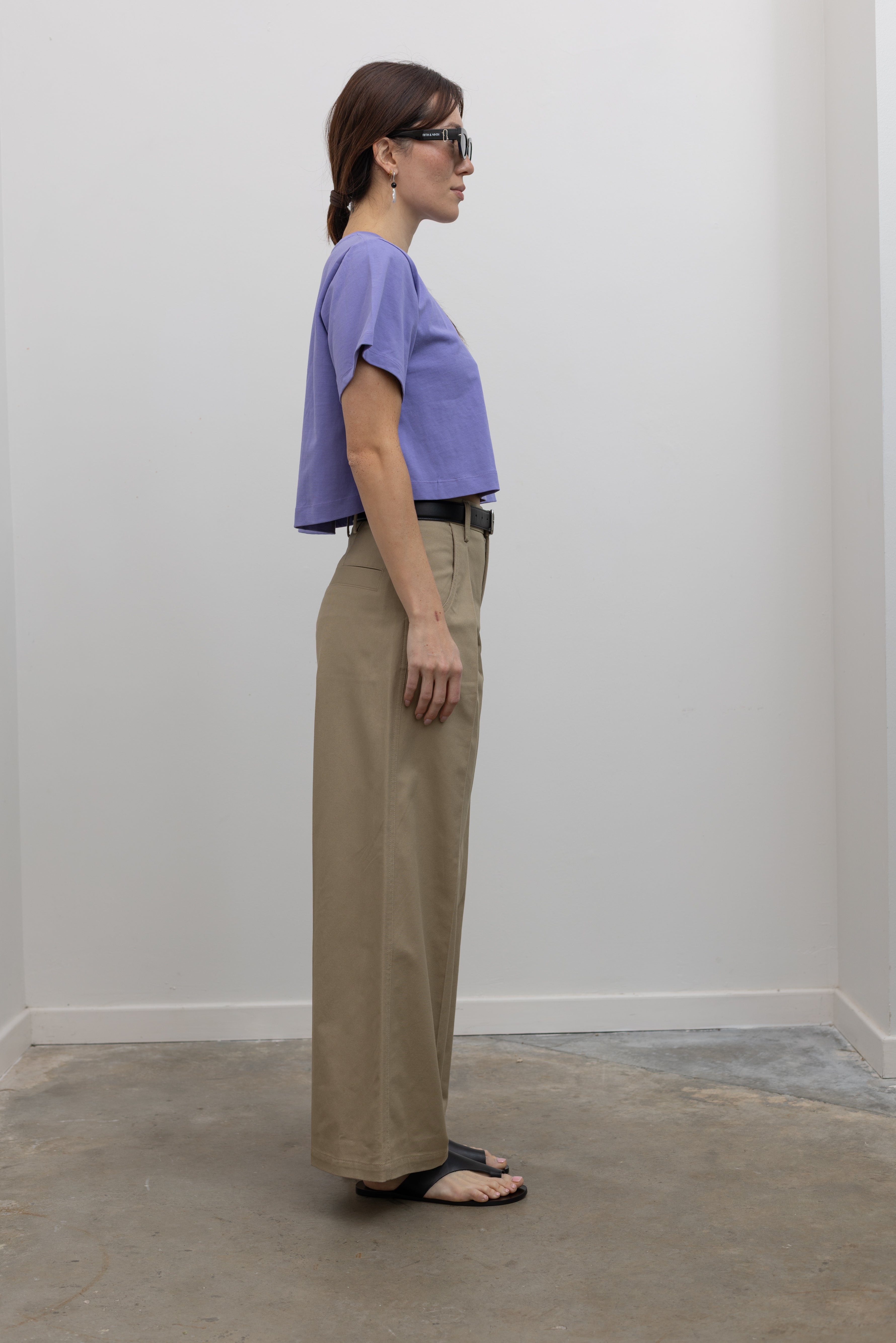 Side view of Woman standing wearing Spring fashion in a pair of Tela Rubicon Sand Cotton Gabardine Trousers and cropped blue tee with sunglasses and sandals at Alas Eius