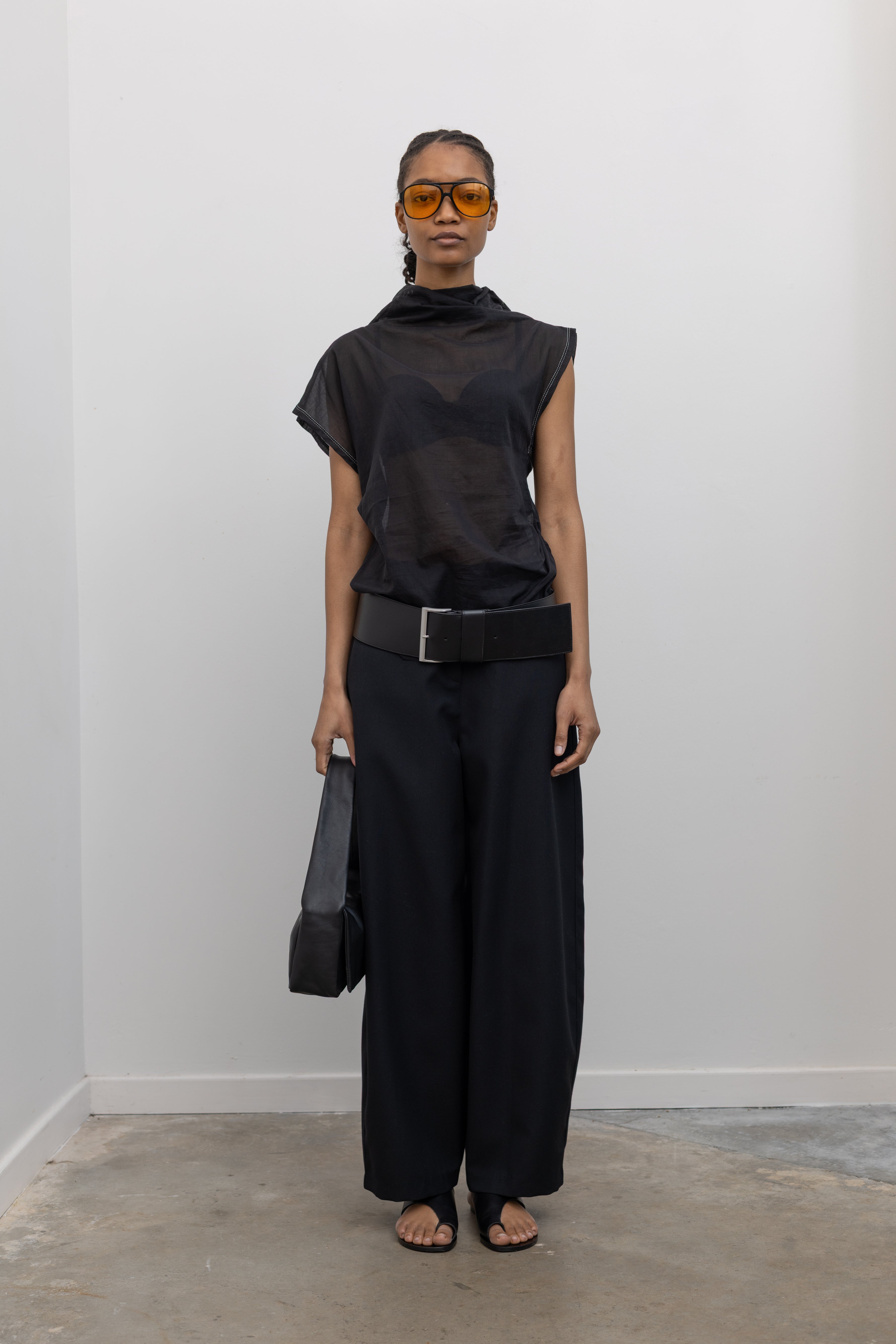 Full length front view of a woman posing wearing chic and feminine spring fashion in a Tela semi-sheer lightweight washed muslin black top featuring a soft draped asymmetrical neckline paired with a classic wide black leather belt on black wide leg trousers at an Alas Eius store. 