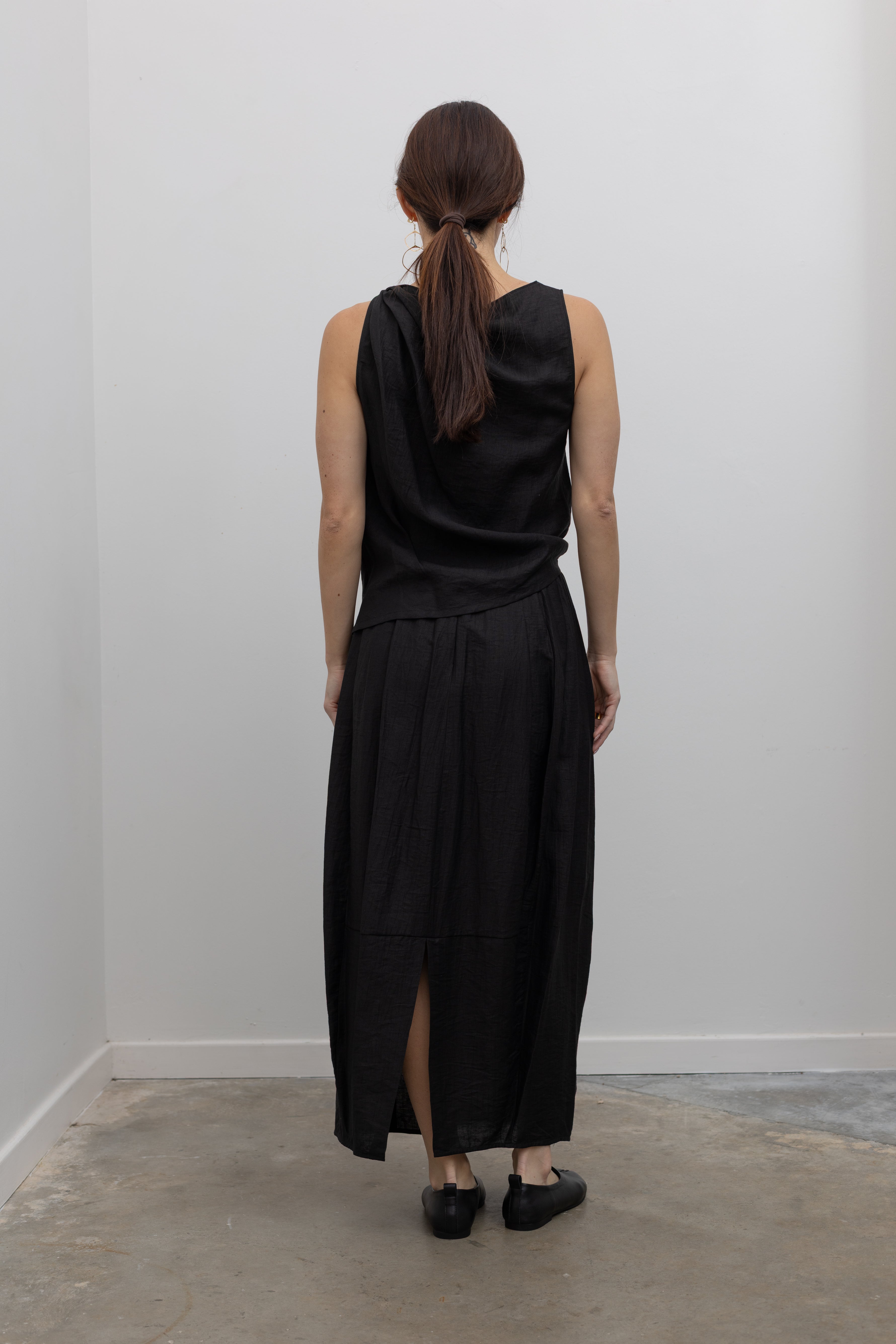 Full back view of a woman standing wearing Spring fashion in an elegantly draped Tela Cody black linen knotted shoulder sleeveless top at Alas Eius. 