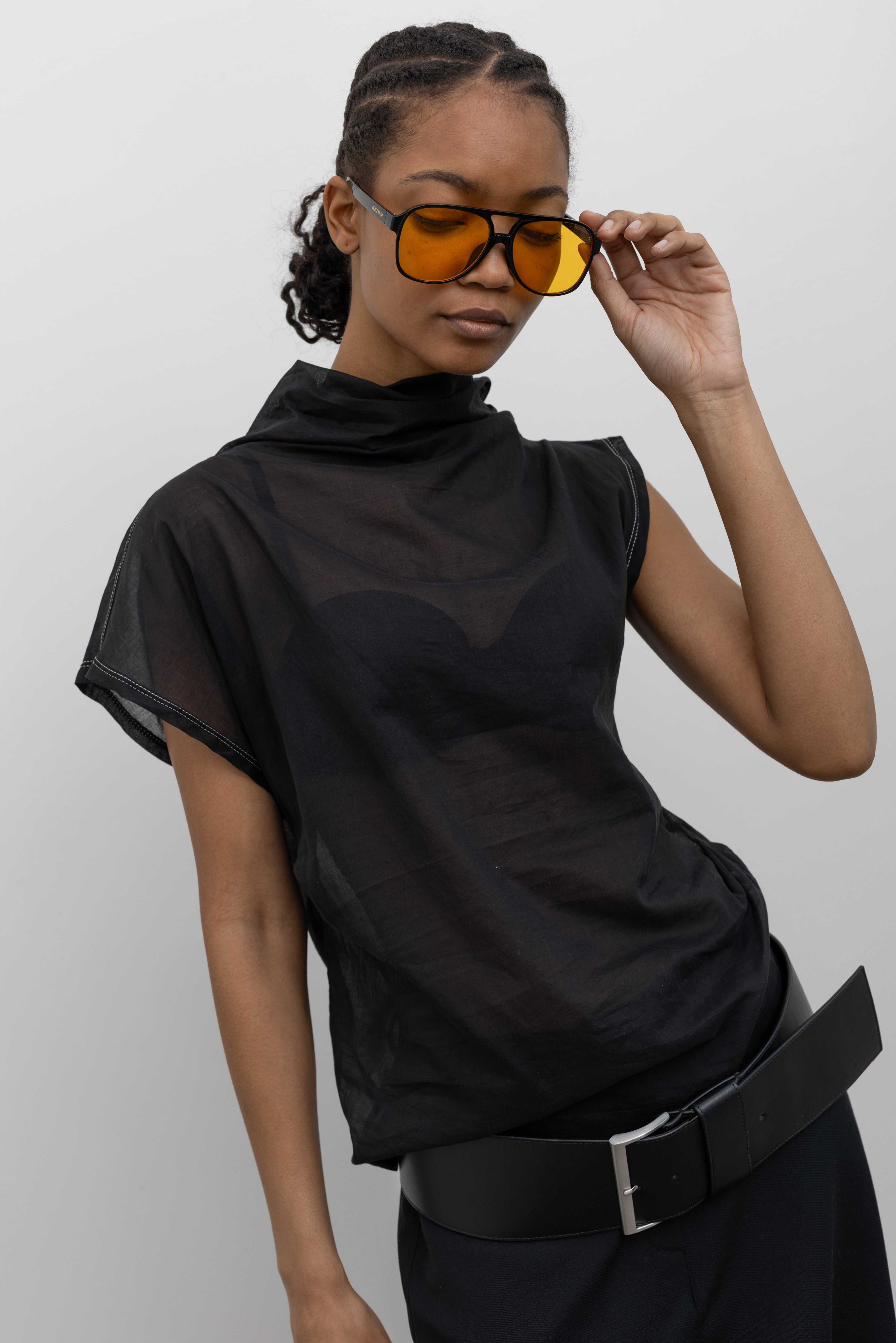 Woman posing wearing chic and feminine spring fashion in a Tela semi-sheer lightweight washed muslin black top featuring a soft draped asymmetrical neckline paired with a classic wide black leather belt at an Alas Eius store. 