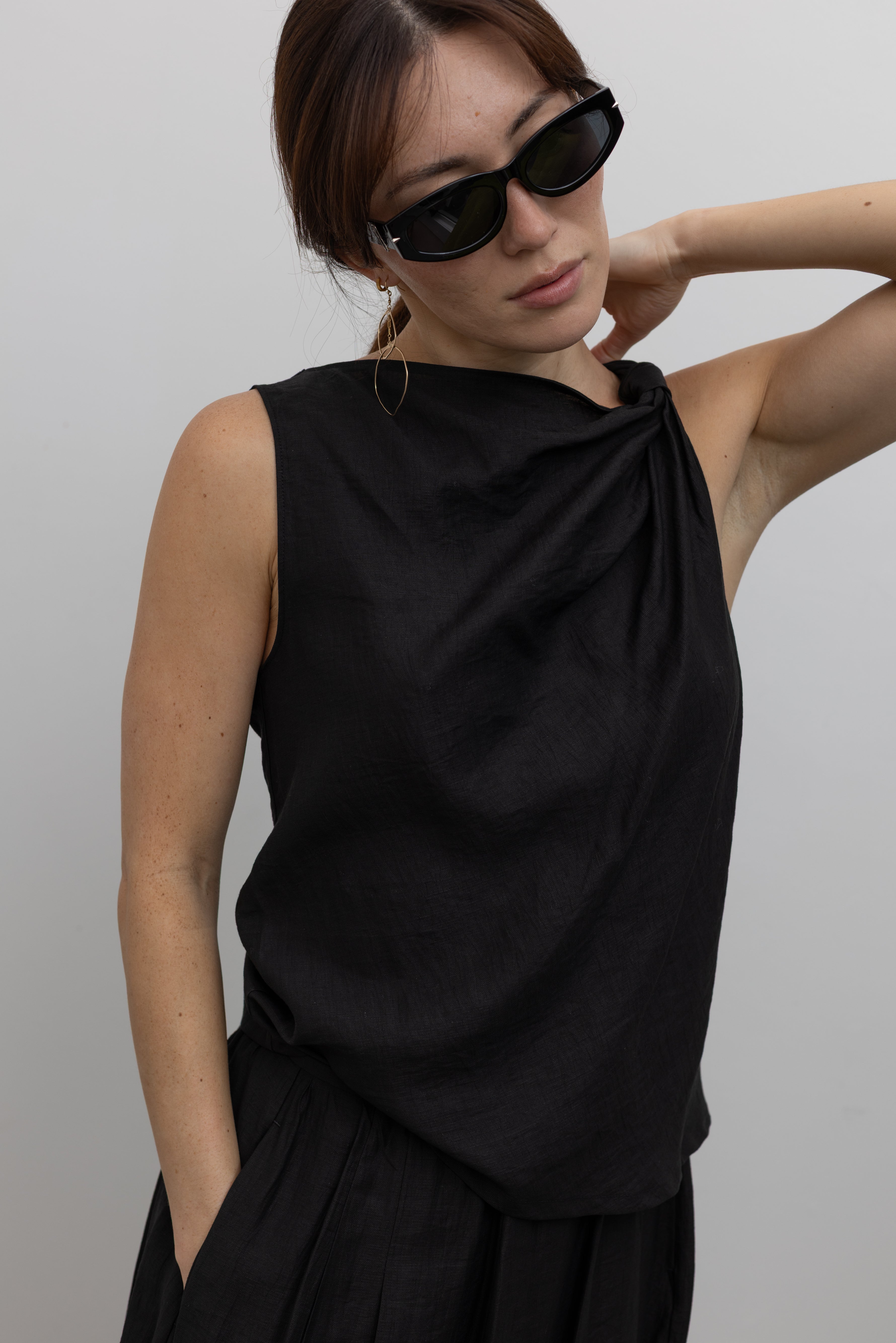 Close-up front view of a woman standing wearing Spring fashion in an elegantly draped Tela Cody black linen knotted shoulder sleeveless top at Alas Eius. 