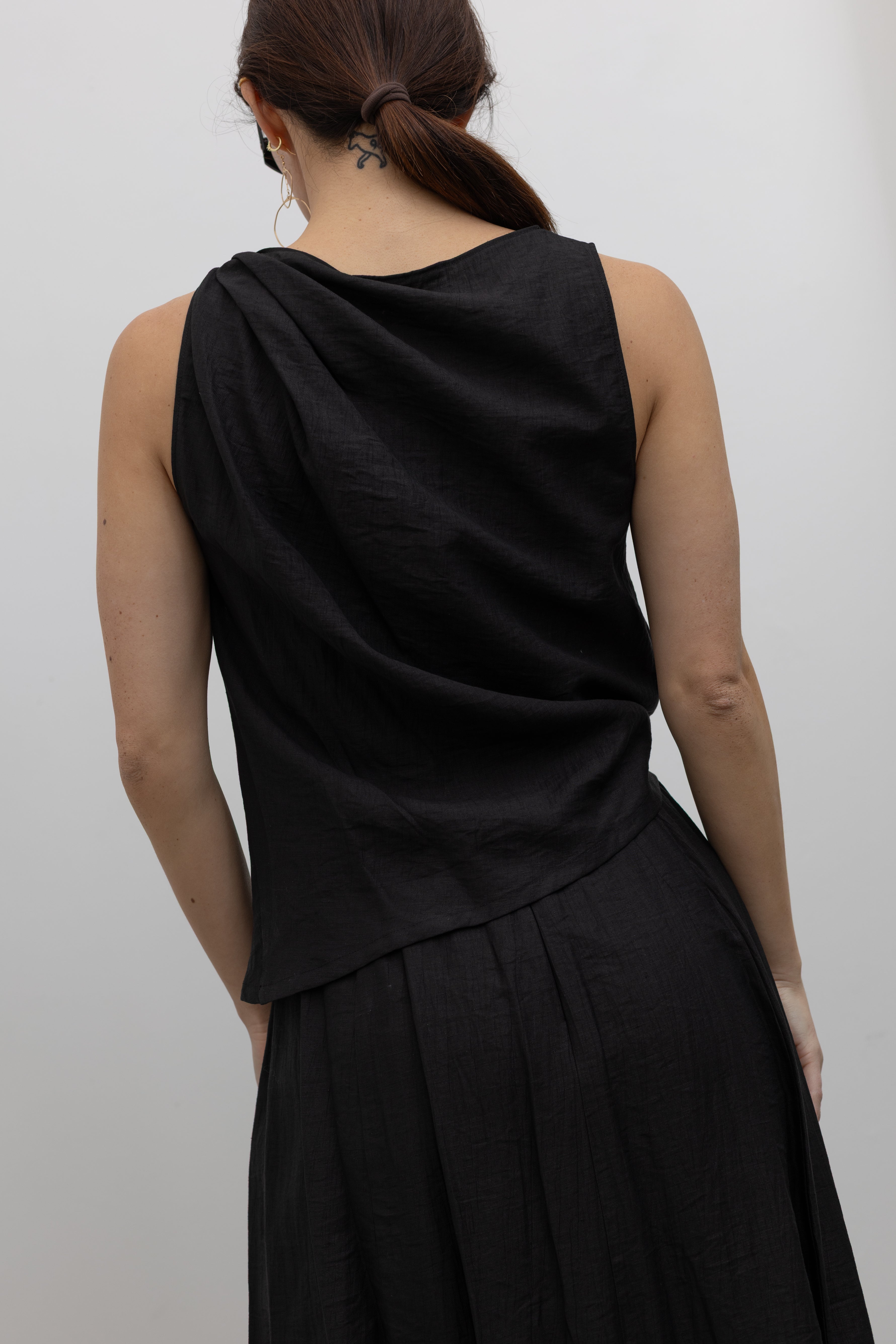 Close-up back view of a woman standing wearing Spring fashion in an elegantly draped Tela Cody black linen knotted shoulder sleeveless top at Alas Eius. 