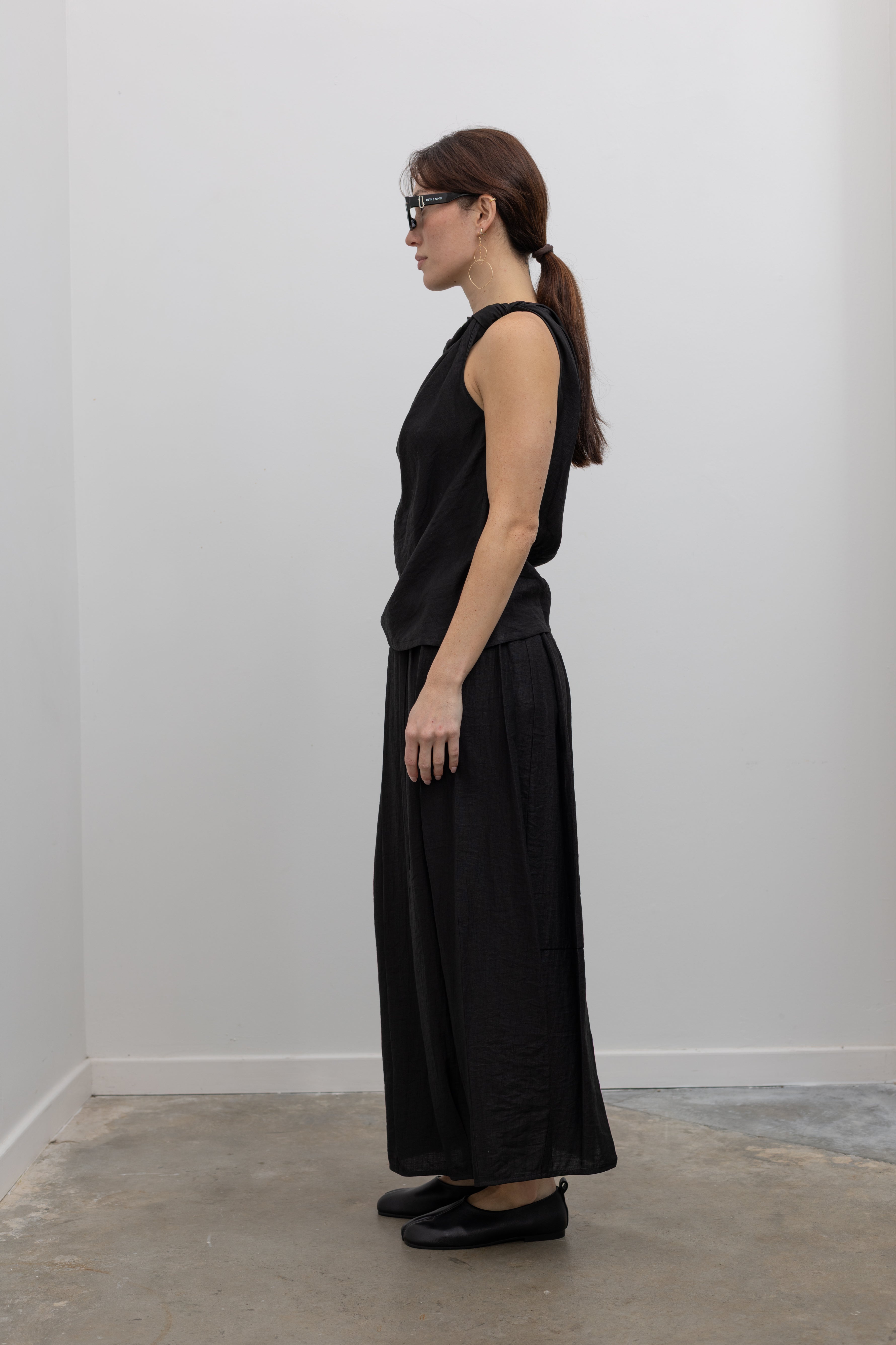 Full side view of a woman standing wearing Spring fashion in a Tela Candito black linen flowy maxi skirt with an elastic waistband and subtle back slit pair with an elegant Tela black linen knotted shoulder draped top at Alas Eius.
