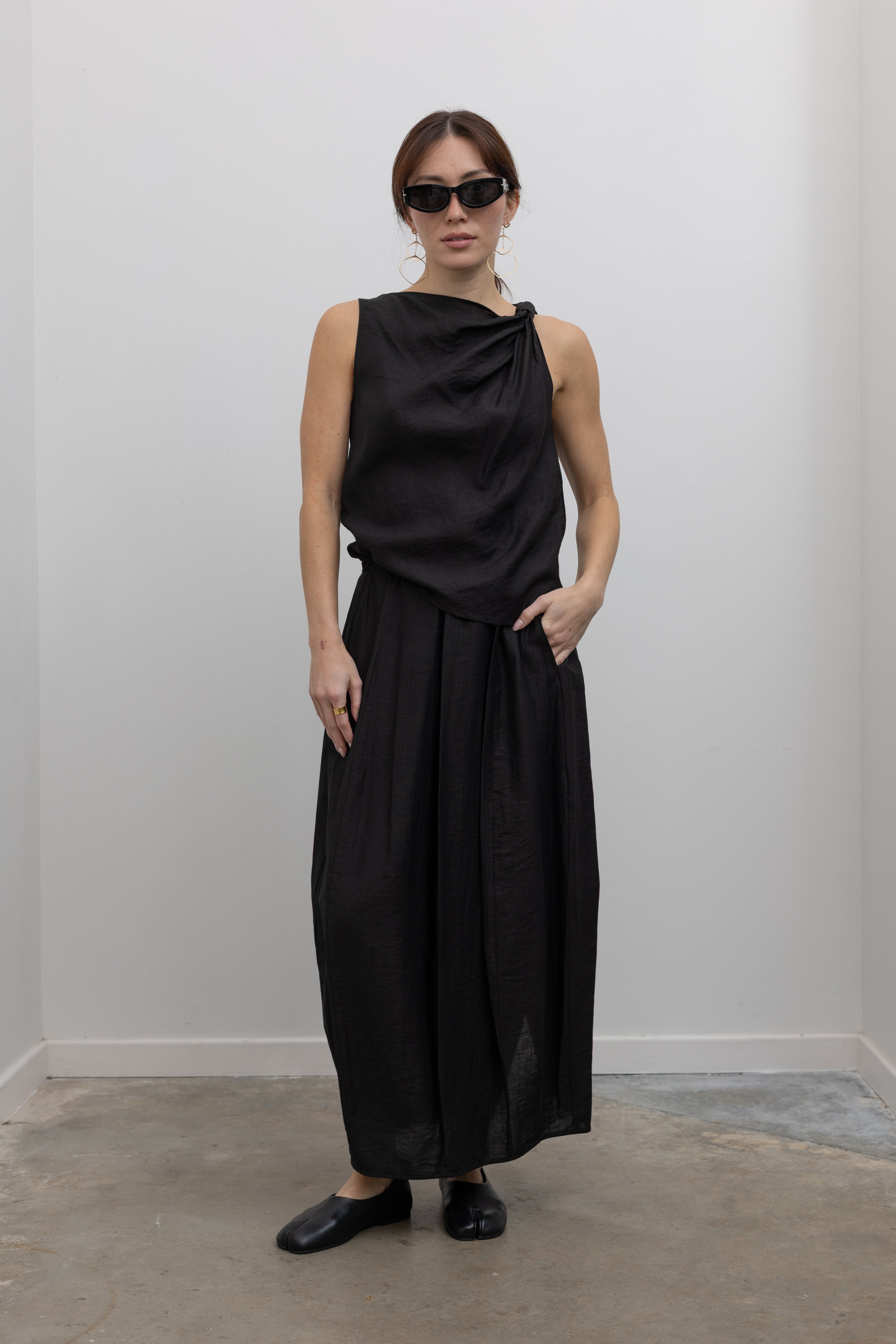 Full front view of a woman standing wearing Spring fashion in a Tela Candito black linen flowy maxi skirt with an elastic waistband, pockets, and subtle back slit pair with an elegant Tela black linen knotted shoulder draped top at Alas Eius.