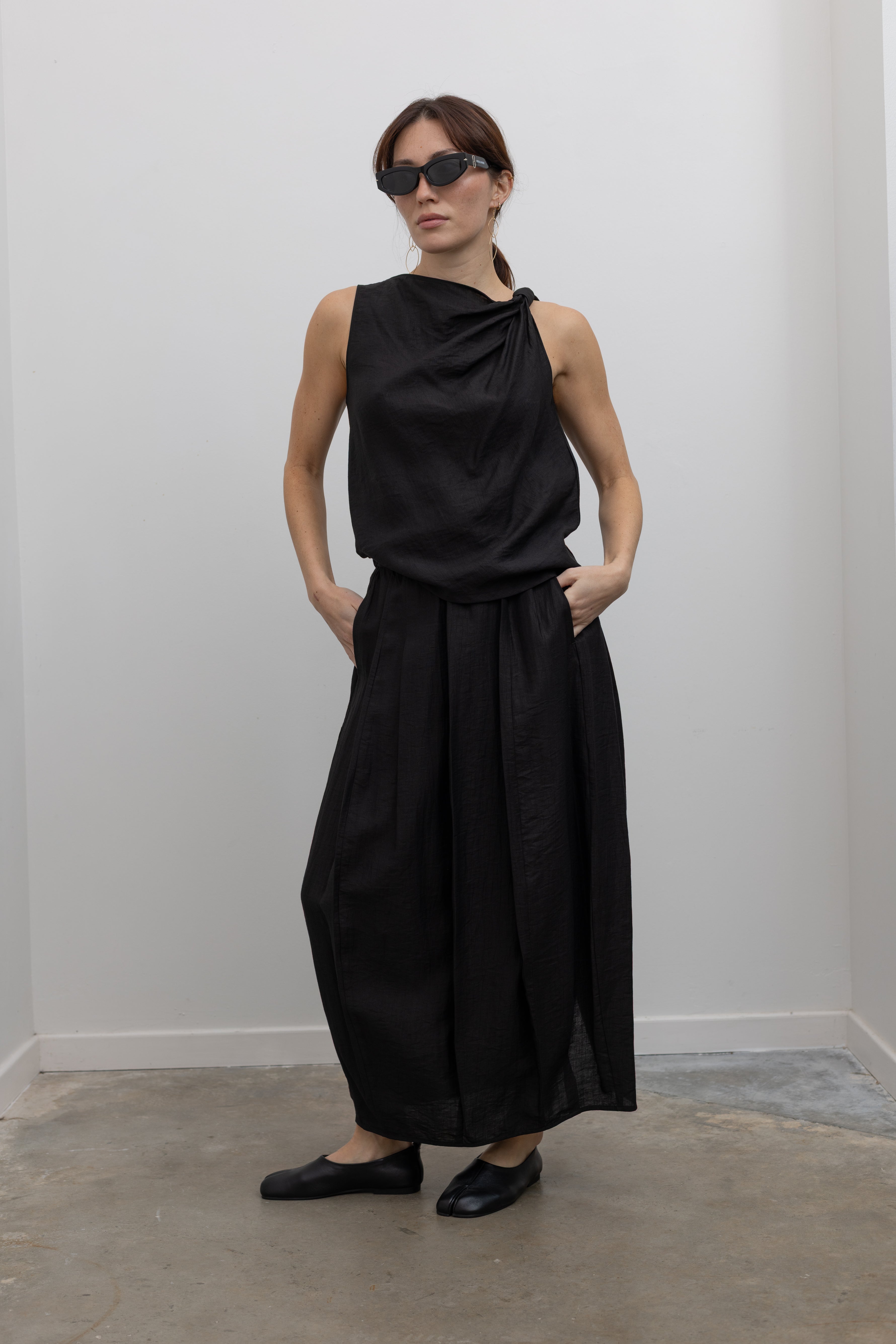 Full front view of a woman posing wearing Spring fashion in a Tela Candito black linen flowy maxi skirt with an elastic waistband and subtle back slit pair with an elegant Tela black linen knotted shoulder draped top at Alas Eius.