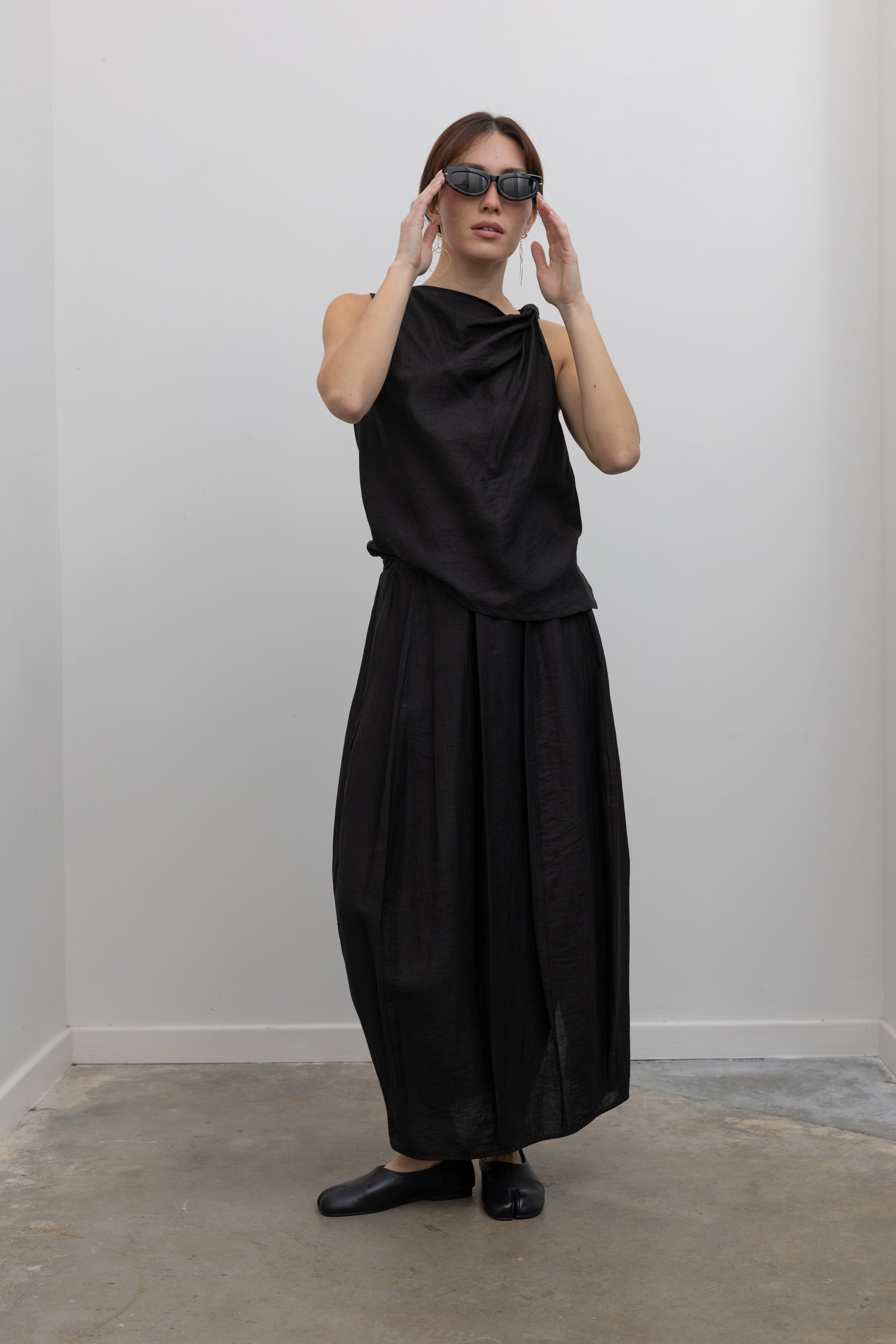 Full front view of a woman posing with sunglasses wearing Spring fashion in a Tela Candito black linen flowy maxi skirt with an elastic waistband and subtle back slit pair with an elegant Tela black linen knotted shoulder draped top at Alas Eius.