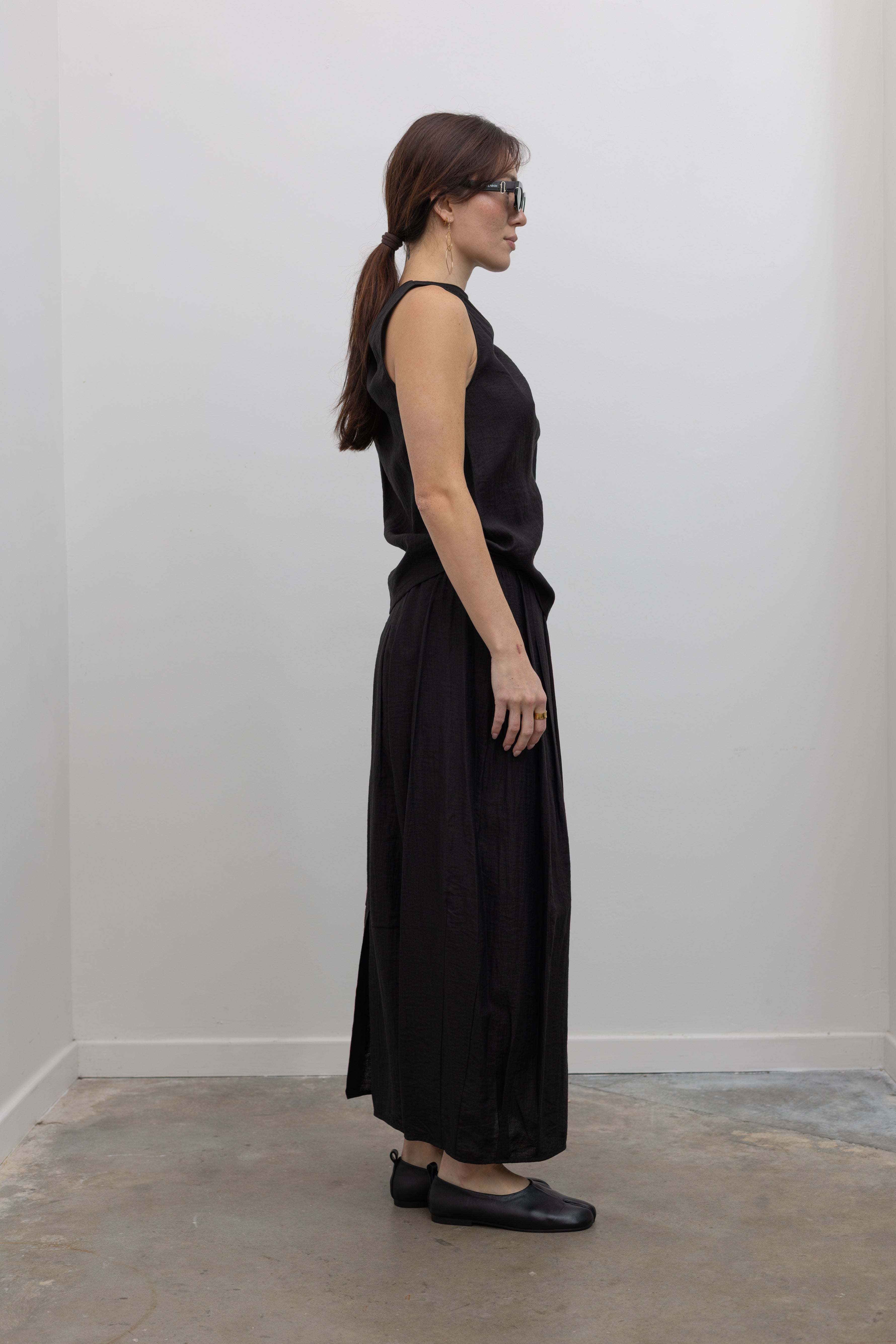 Side view of a woman standing wearing Spring fashion in an elegantly draped Tela Cody black linen knotted shoulder sleeveless top paired with a black pleated ankle length skirt and black leather flats at Alas Eius. 