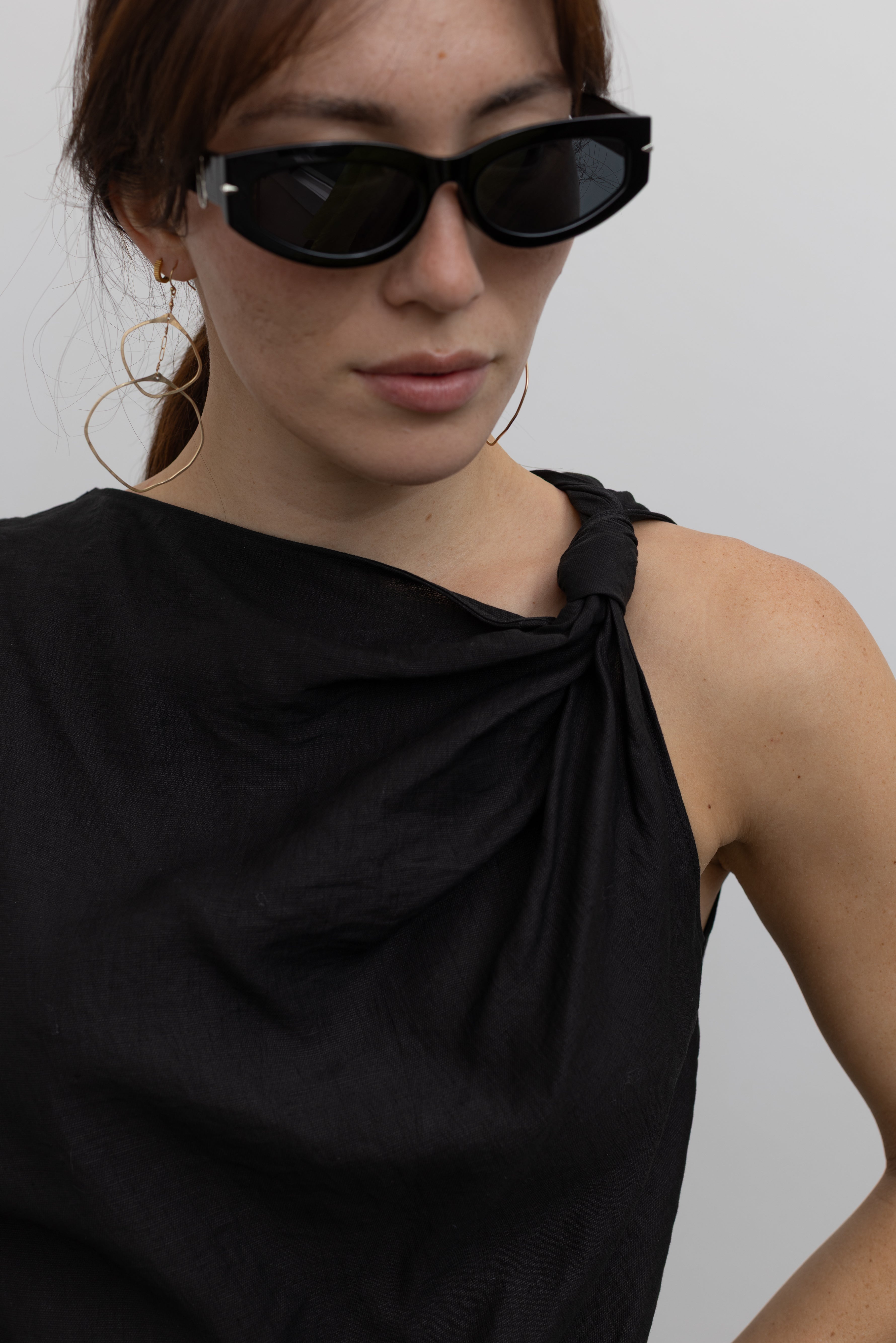 Close-up front view of a woman standing wearing Spring fashion in an elegantly draped Tela Cody black linen knotted shoulder sleeveless top at Alas Eius. 