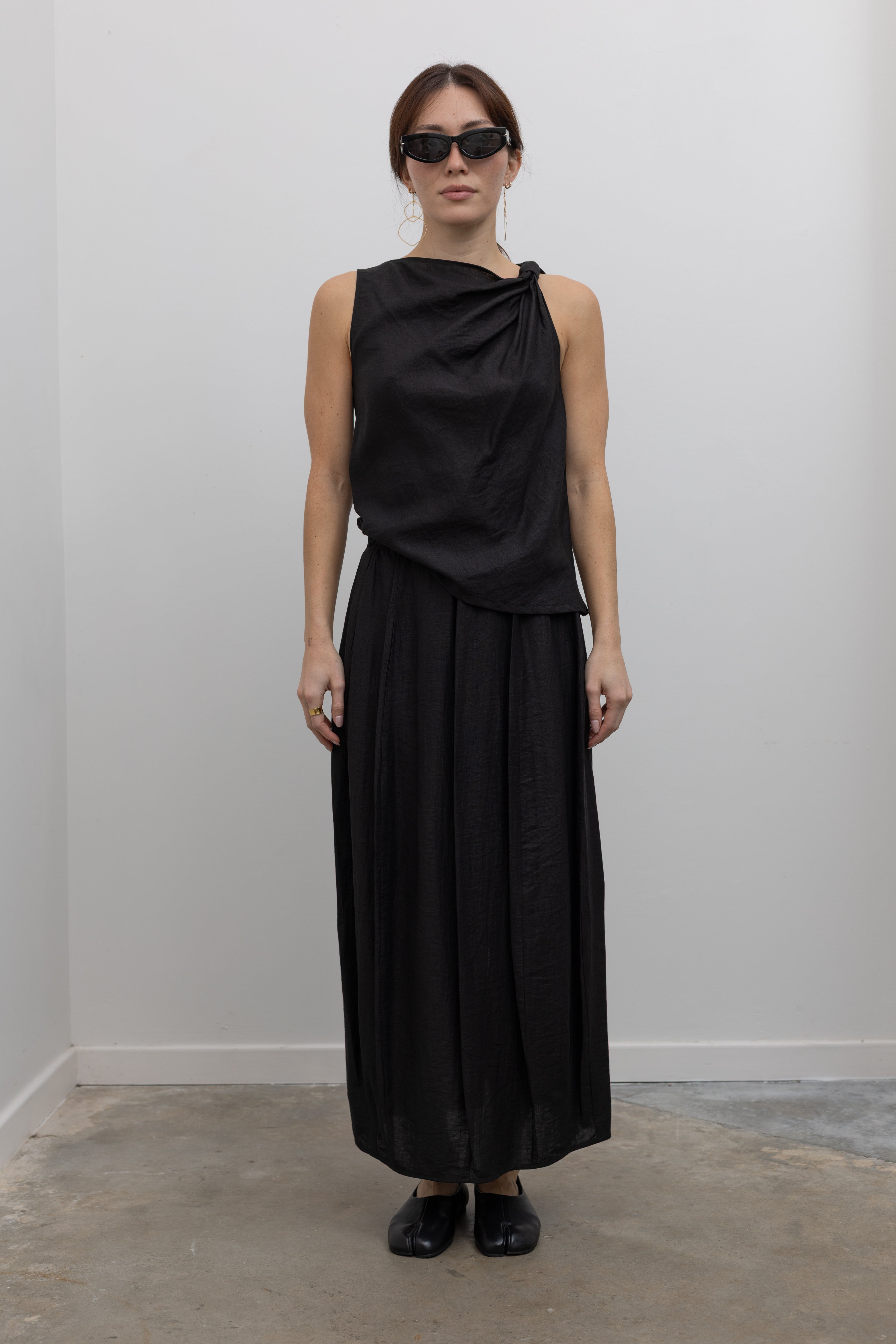 Full front view of a woman standing wearing Spring fashion in an elegant and flattering Tela Cody black linen knotted shoulder sleeveless top paired with an ankle length pleated maxi skirt and black leather flats at Alas Eius. 