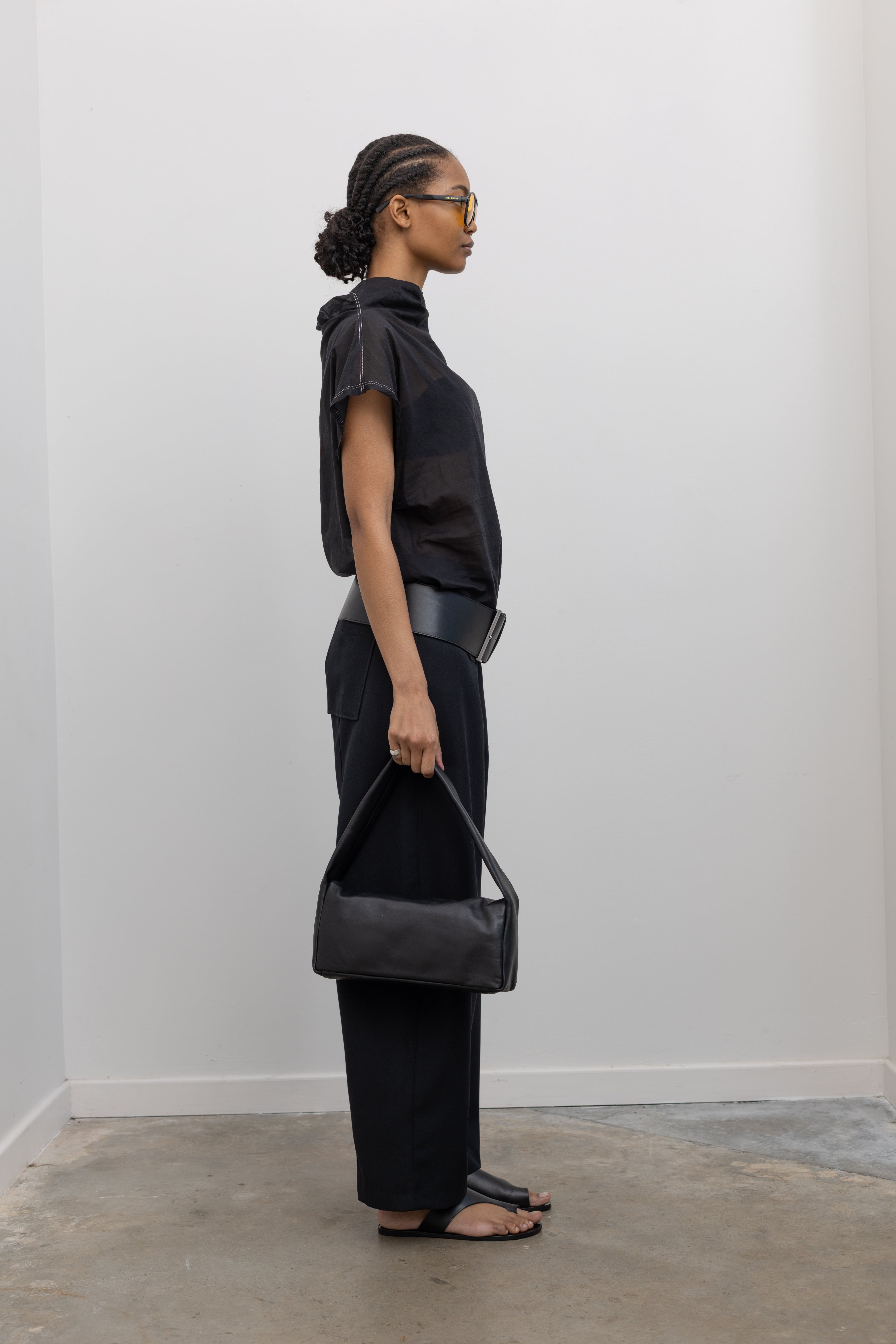 Full length side view of a woman posing wearing chic and feminine spring fashion in a Tela semi-sheer lightweight washed muslin black top featuring a soft draped asymmetrical neckline paired with a classic wide black leather belt on black wide leg trousers at an Alas Eius store. 