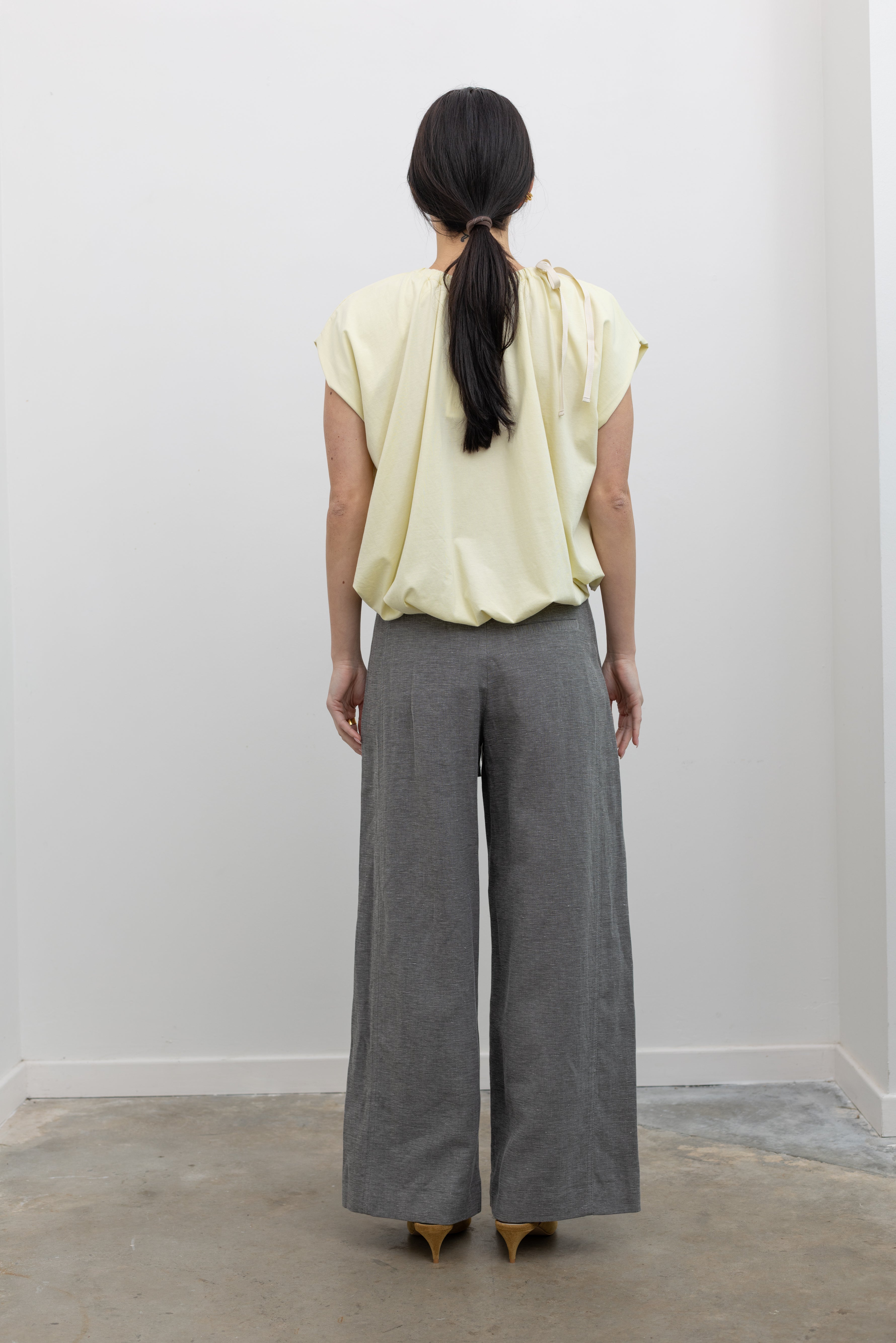 Full length back view of a woman standing wearing modern minimalist spring fashion in a Tela Light Yellow Gathered Neck Bubble Hem Top, Tela Lucio light gray drawstring pants featuring a wide leg and side seam pockets, and Tela Feng Mustard Leather Heeled Sandals at an Alas Eius store. 