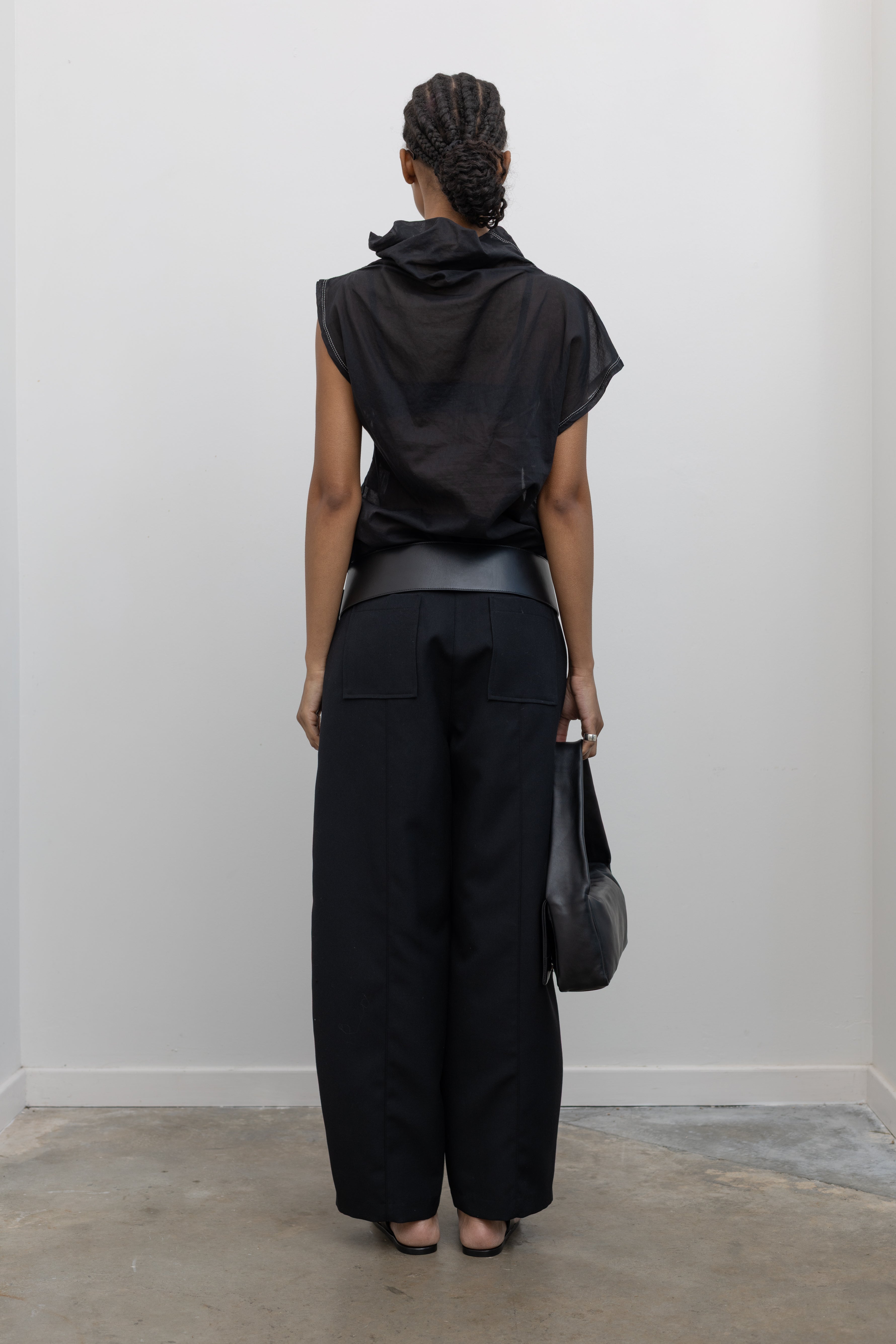Full length back view of a woman wearing contemporary spring fashion in a Tela semi-sheer lightweight washed muslin black top featuring a soft draped asymmetrical neckline paired with a classic wide black leather belt on black wide leg trousers at an Alas Eius store. 