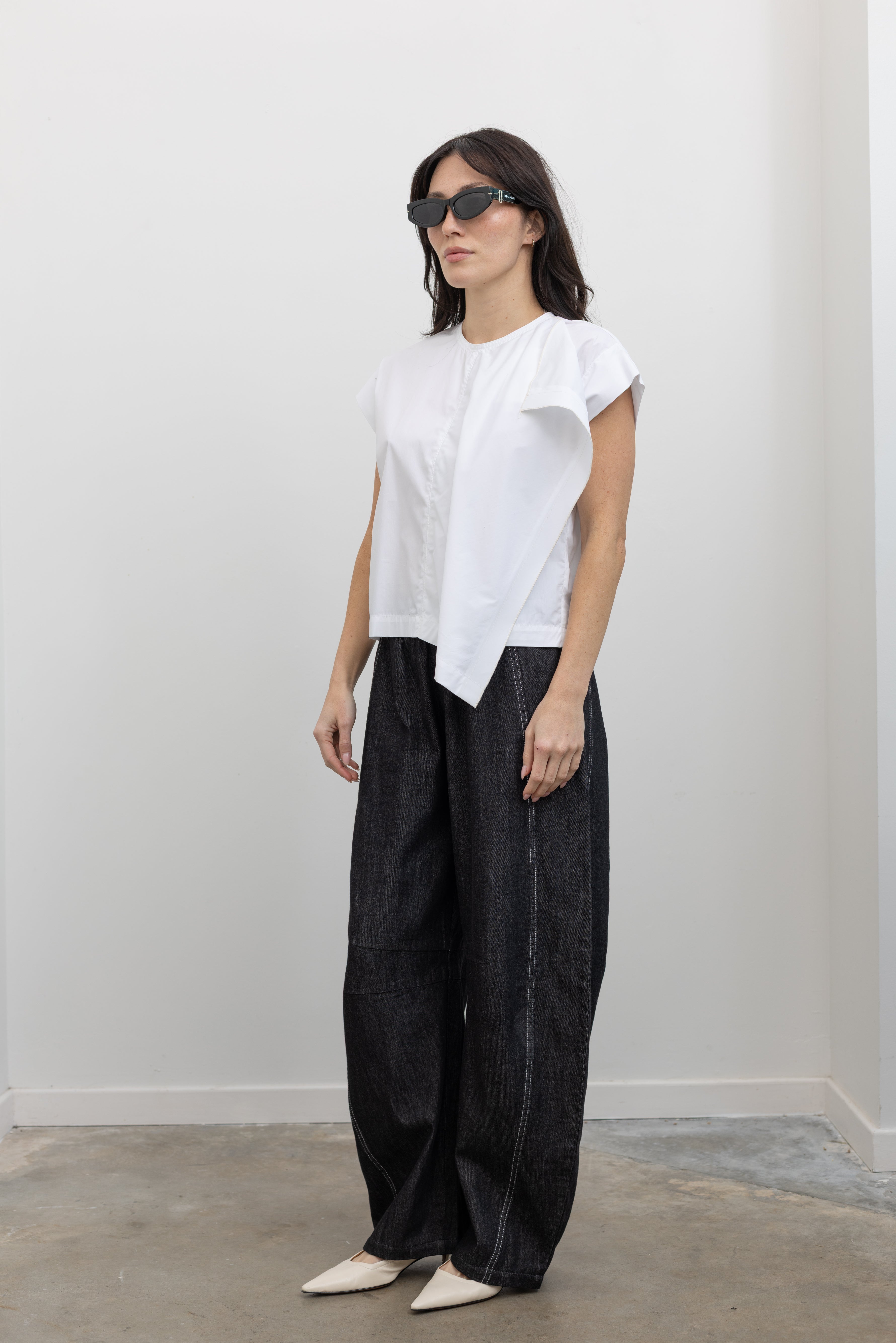 Full length 3/4 view of a woman wearing modern minimalist spring fashion in a Tela brand white Livia shirt featuring a front pleat that creates an intentional asymmetrical draped panel, crew neckline and relaxed cap sleeves paired with oversized baggy barrel leg pants at at an Alas Eius store. 