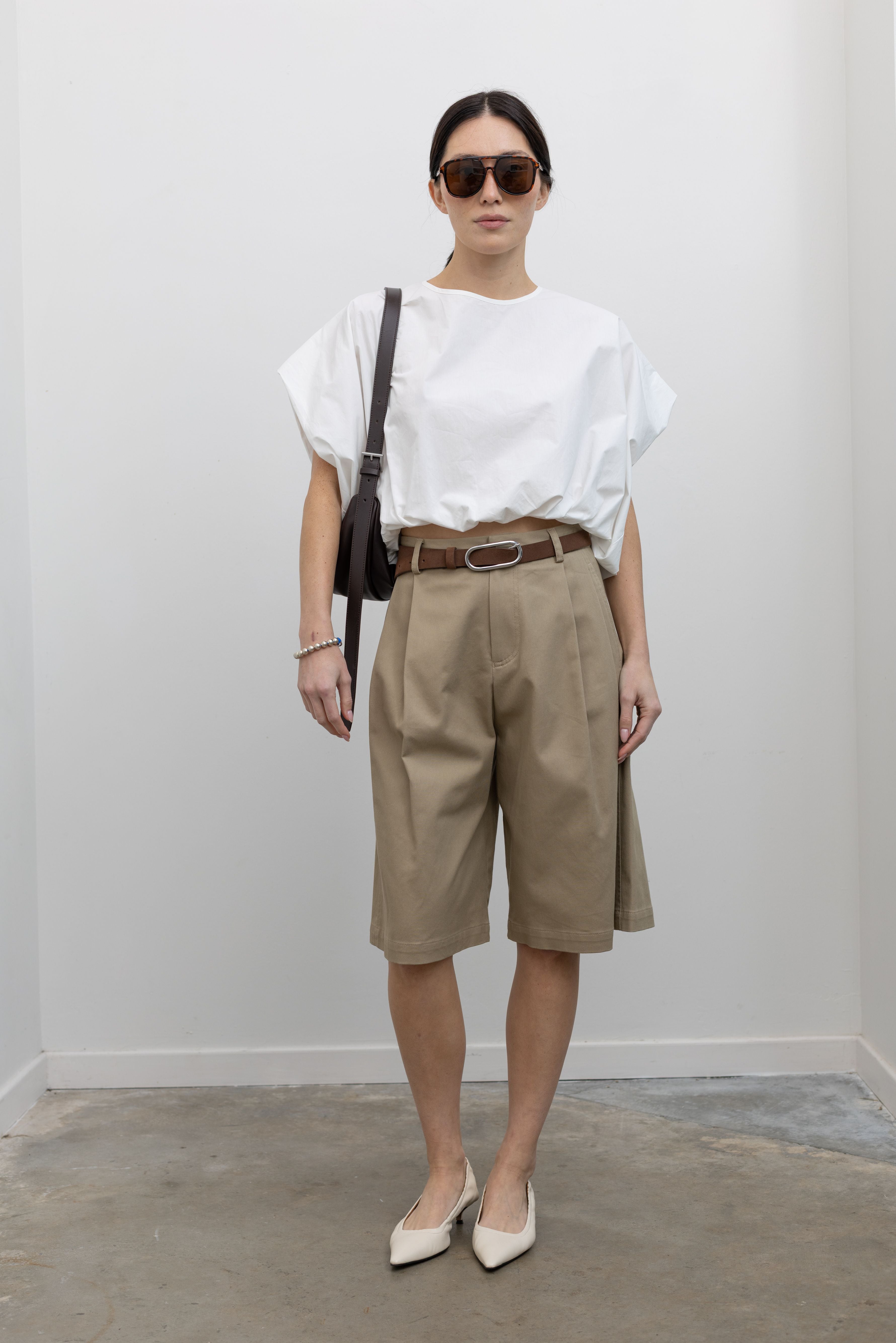 Full length front view of a woman posing wearing an elevated and chic outfit in the Tela sand cotton gabardine Bermuda long short featuring a front pleat and side seam pockets paired with a classic brown leather belt, a crisp white top, a St. Agni leather bag, and white leather pointed toe kitten heels at an Alas Eius store. 