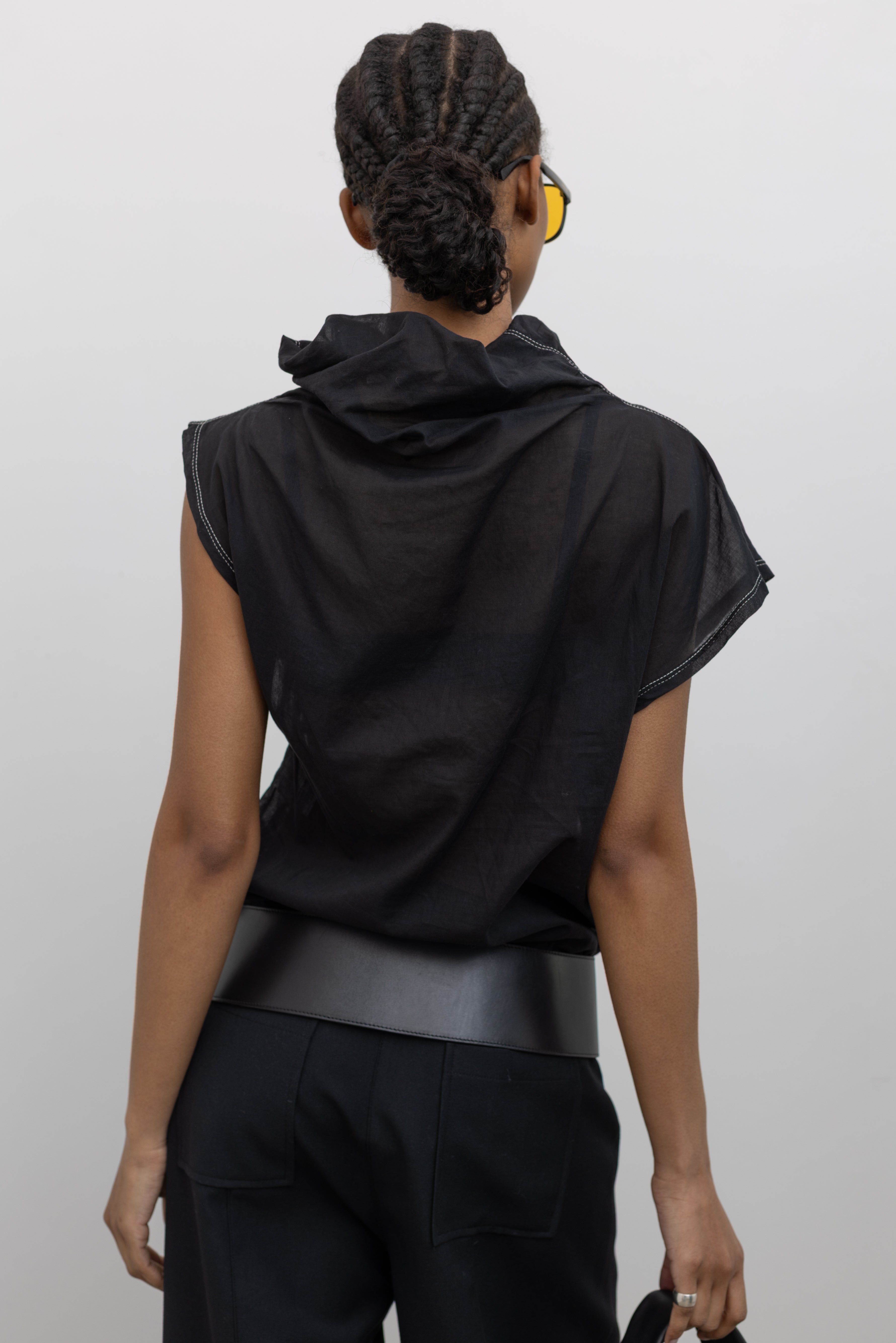 Close-up back view of a woman posing wearing a unique Tela brand semi-sheer lightweight washed muslin black top featuring a soft draped asymmetrical neckline paired with a classic wide black leather belt on black wide leg trousers at an Alas Eius store. 