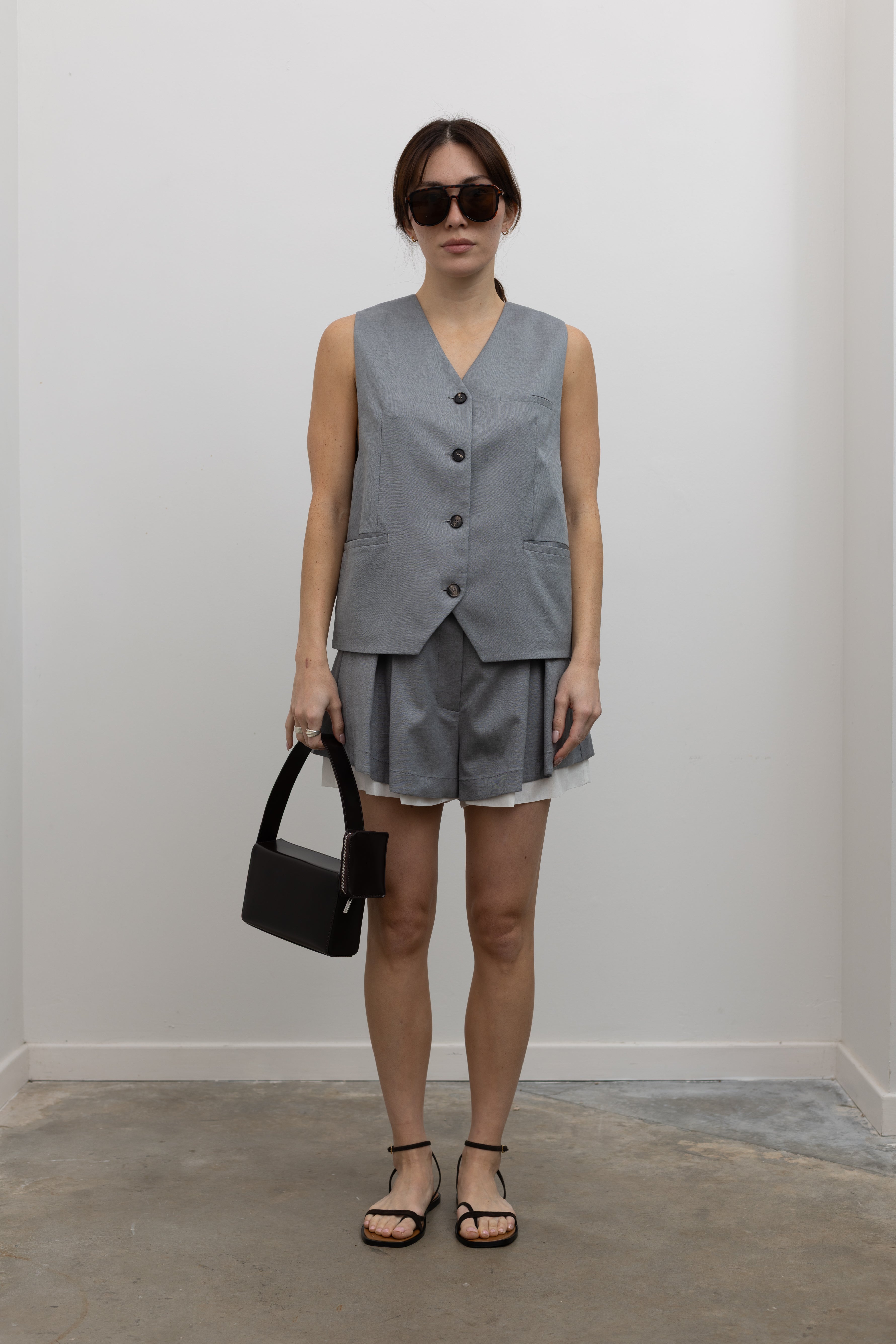 Full length front view of a woman wearing Spring fashion in a sophisticated and refined Tela Gabriel Grey Cool Wool Button Down Vest paired with Tela grey wool layered shorts and black leather strappy sandals at Alas Eius.
