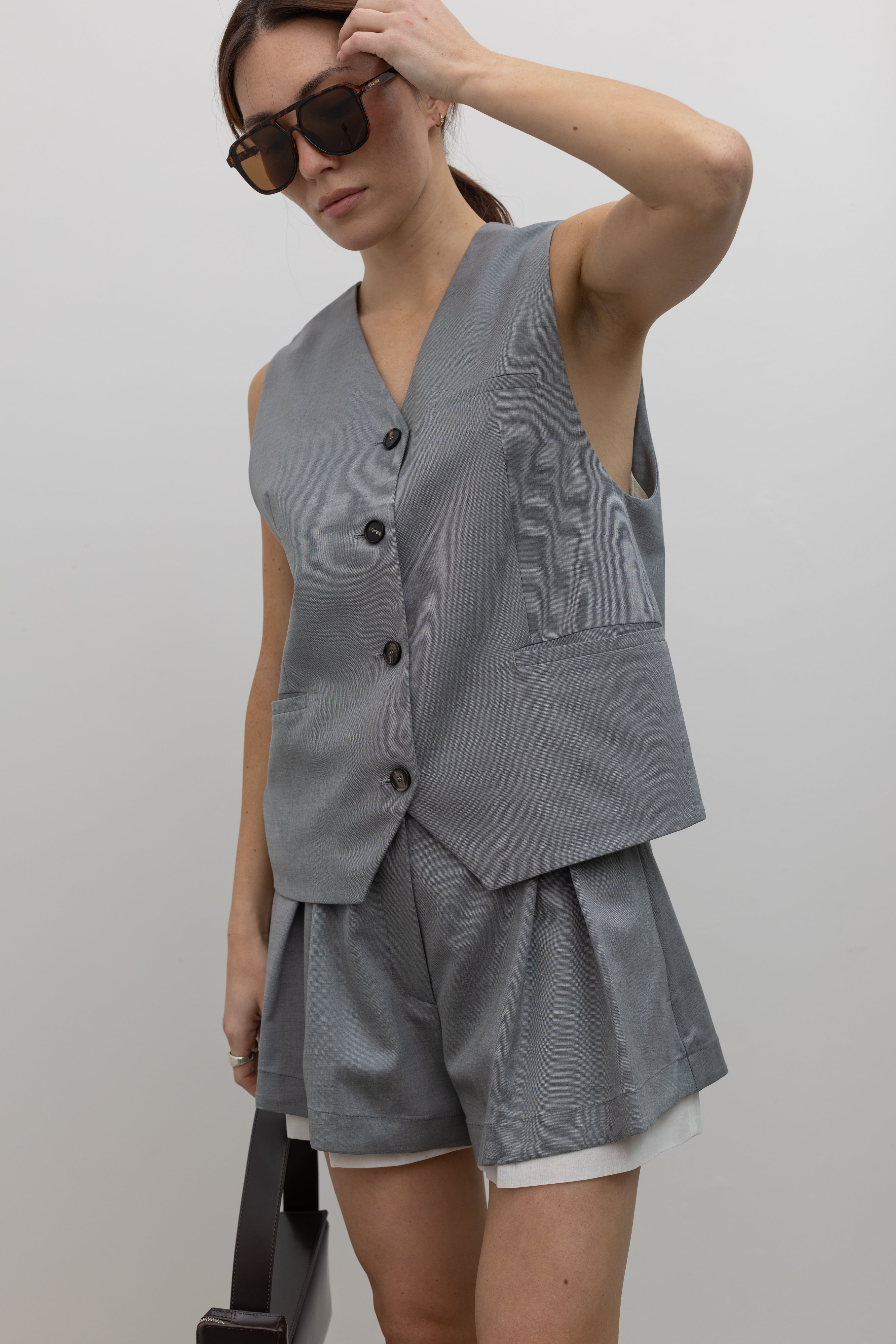 Close-up view of a woman wearing Spring fashion in the structured silhouette of a Tela Gabriel Grey Cool Wool Button Down Vest with Tela grey wool layered shorts and sunglasses at Alas Eius. 