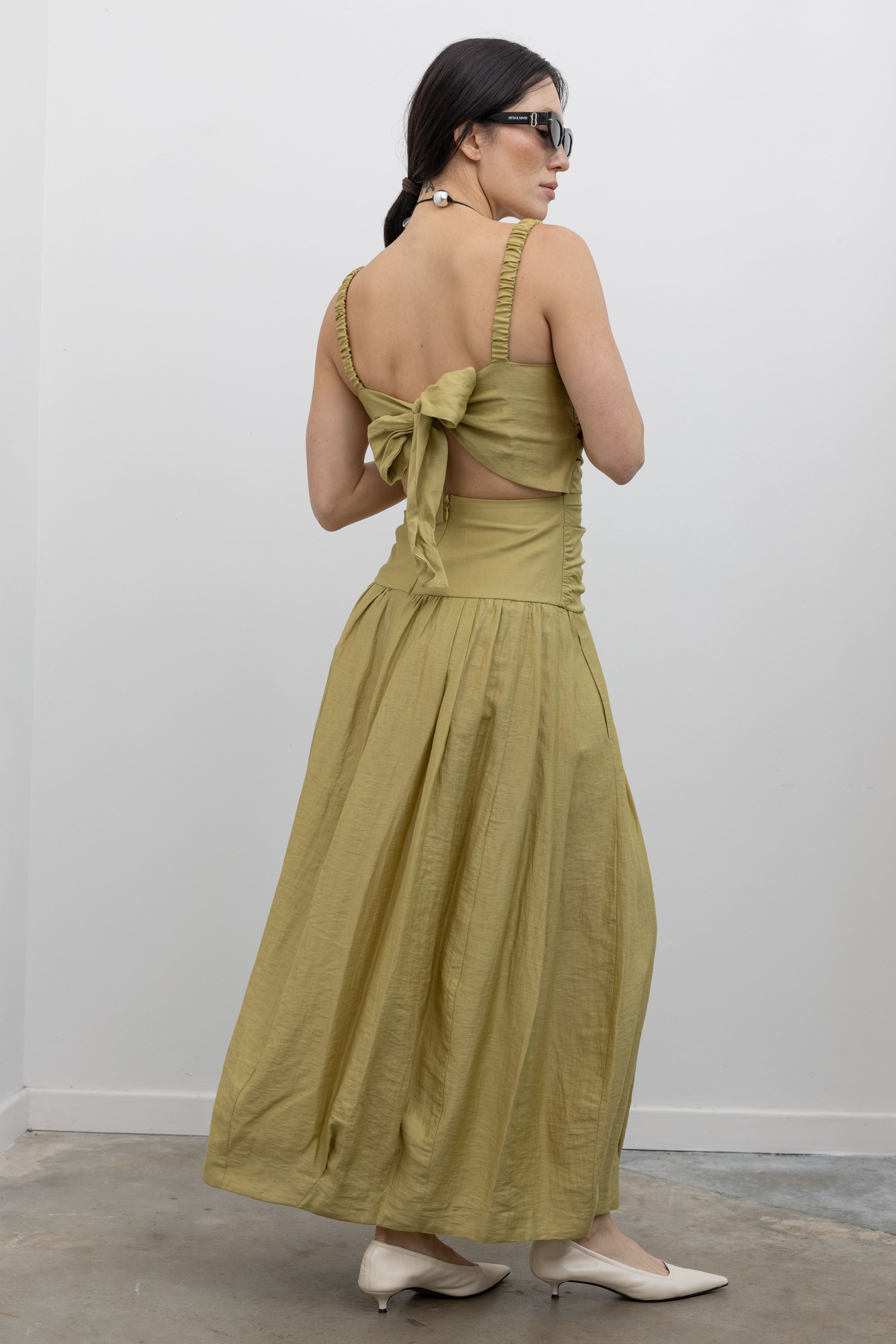 Full length back view of a woman standing wearing modern and elegant spring fashion in a Tela label linen blend pistachio green maxi dress with flattering ruching, delicate narrow shoulder straps, and a striking back cut-out detail with a bow closure paired with white leather slingback kitten heels at an Alas Eius store. 