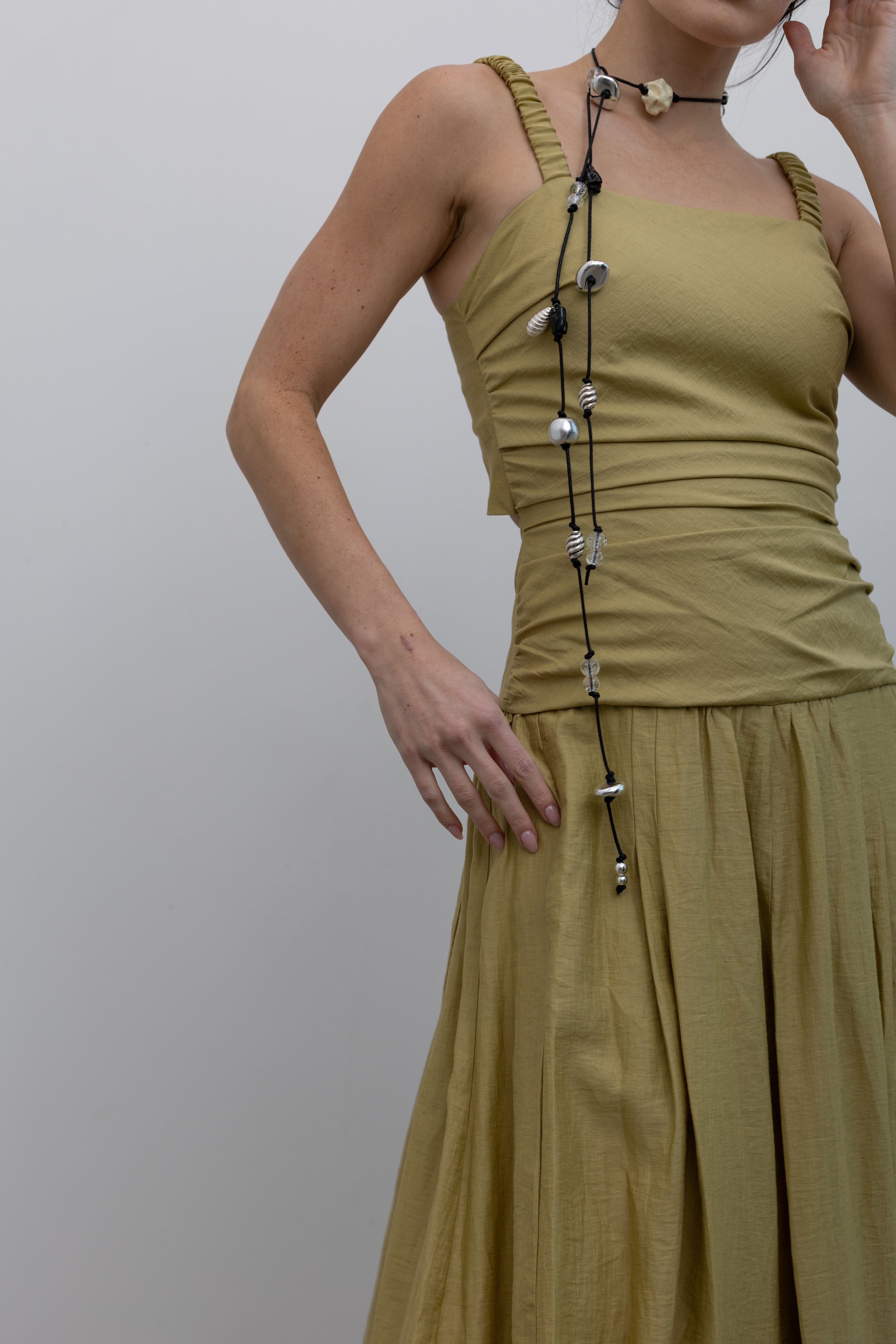 Close-up front view of a woman standing wearing modern and elegant spring fashion in a Tela label linen blend pistachio green maxi dress with flattering ruching, delicate narrow shoulder straps, and a striking back cut-out detail with a bow closure paired with white leather slingback kitten heels at an Alas Eius store. 