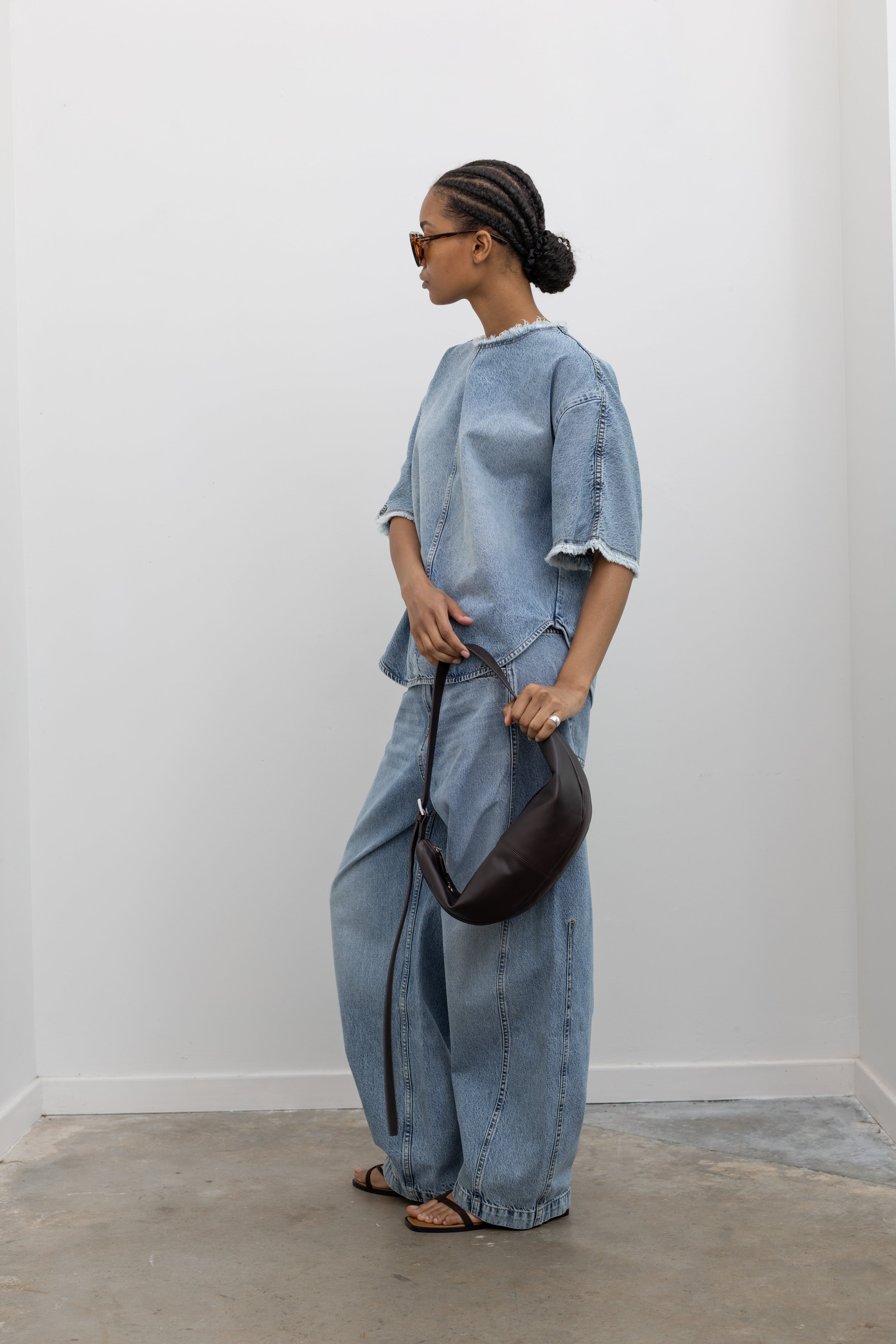 Full length 3/4 view of a Woman wearing modern spring streetwear fashion in a Tela Light Blue Denim Oversized Short Sleeve Shirt featuring  a clean light-blue wash, distinct raw-edge detailing, and a minimalist back neck closure paired with light-blue wash denim jeans at Alas Eius. 