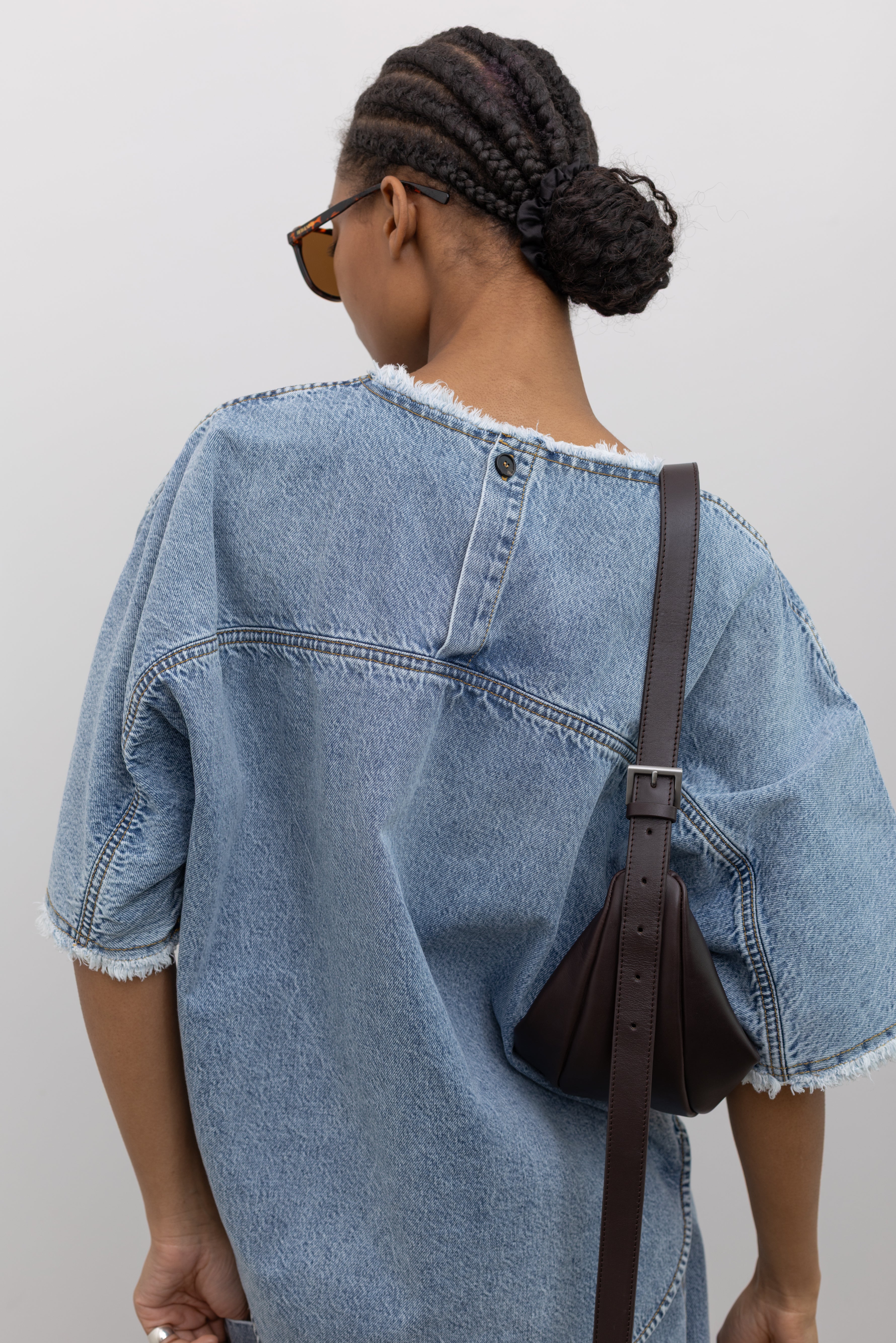 Close-up back view of a Woman wearing elevated spring fashion in a Tela Light Blue Denim Oversized Short Sleeve Shirt featuring a clean light-blue wash, distinct raw-edge detailing, and a minimalist back neck closure paired with a brown leather St. Agni bag at Alas Eius. 