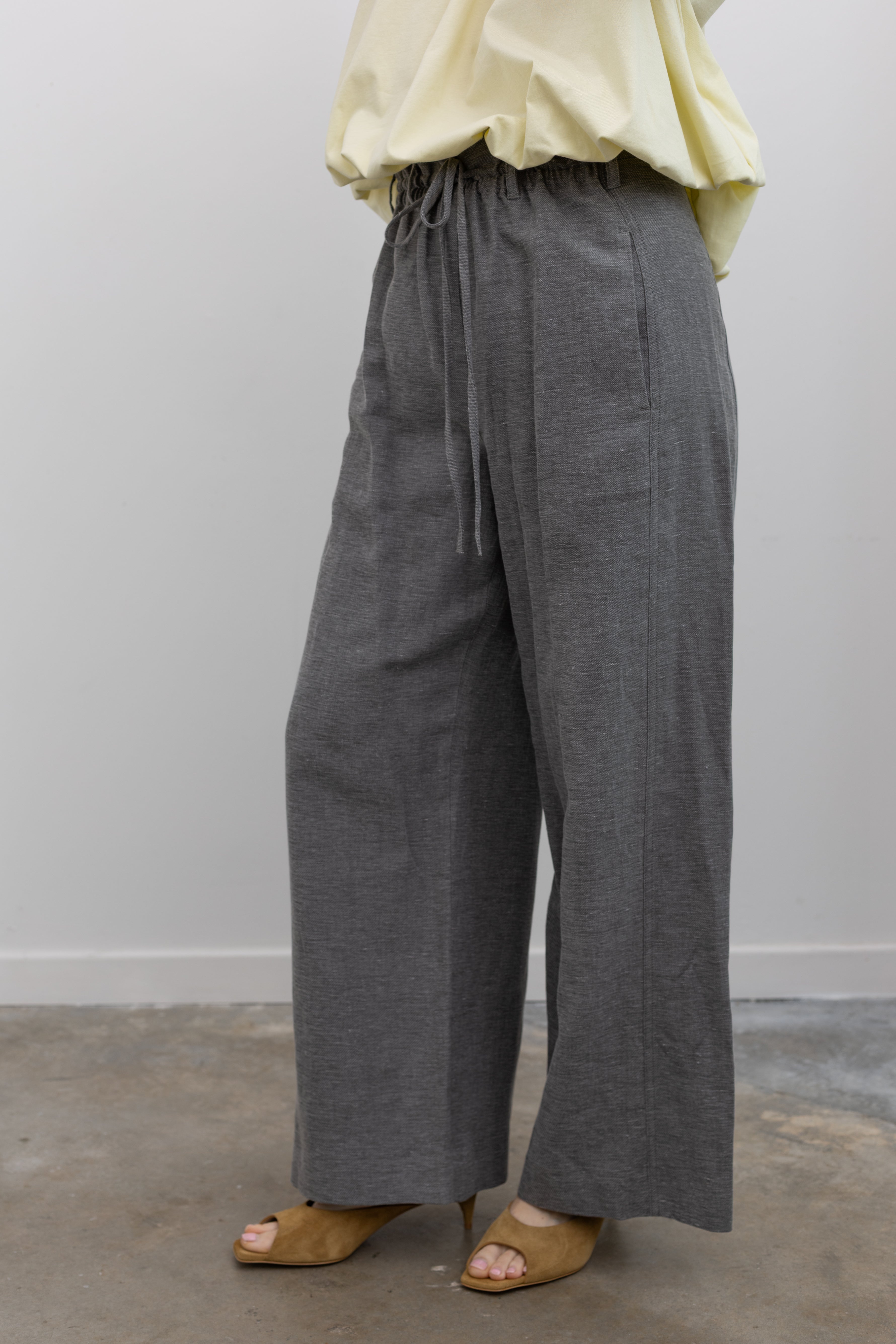 Close-up product view of a woman wearing modern and versatile spring fashion in the Tela Lucio light gray drawstring pants featuring a wide leg and side seam pockets, and Tela Feng Mustard Leather Heeled Sandals at an Alas Eius store. 