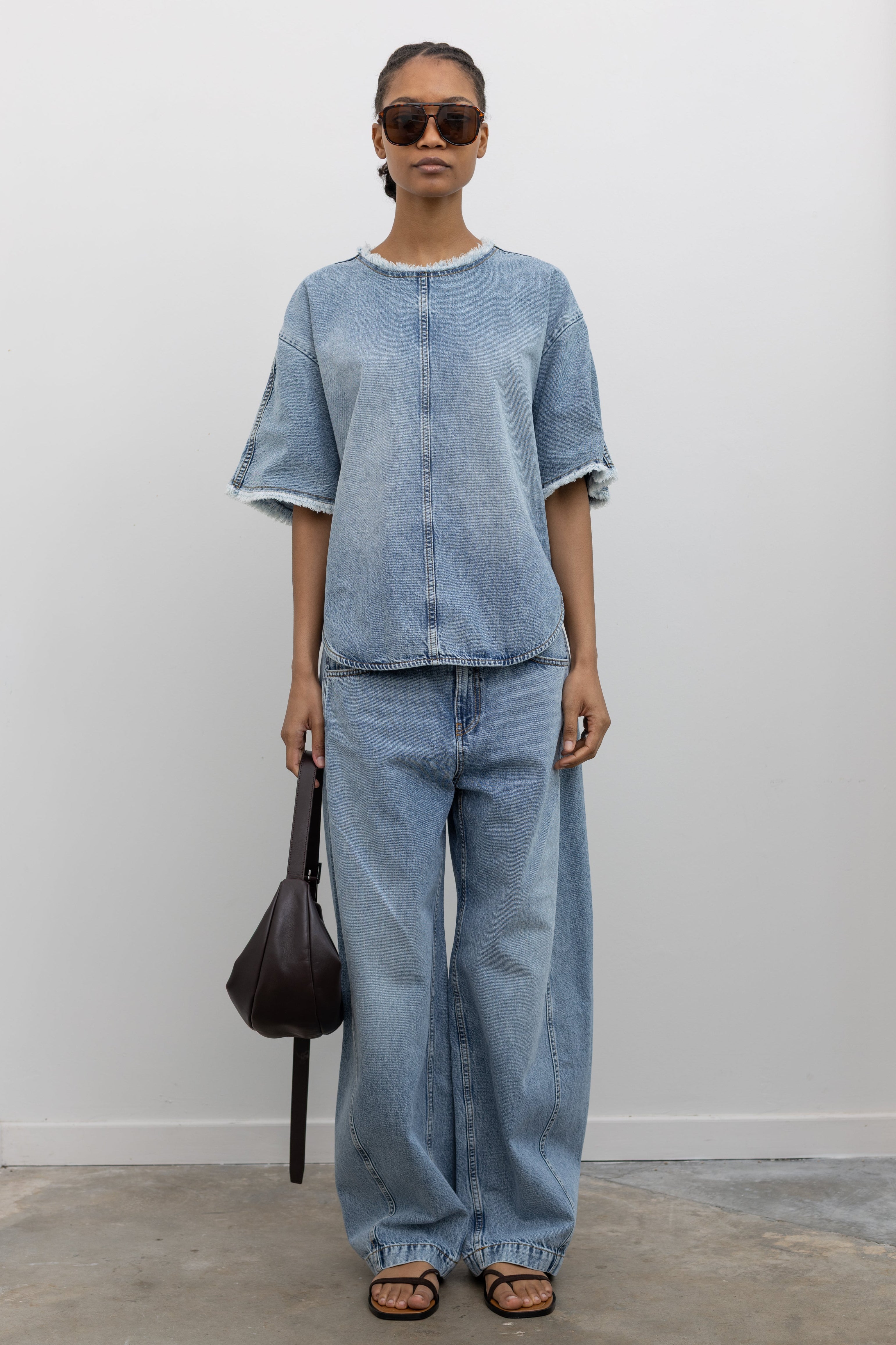 Full length front view of a Woman wearing modern spring streetwear fashion in a Tela Light Blue Denim Oversized Short Sleeve Shirt featuring a clean light-blue wash, distinct raw-edge detailing, and a minimalist back neck closure paired with light-blue wash denim jeans and a leather St. Agni bag at an Alas Eius store. 