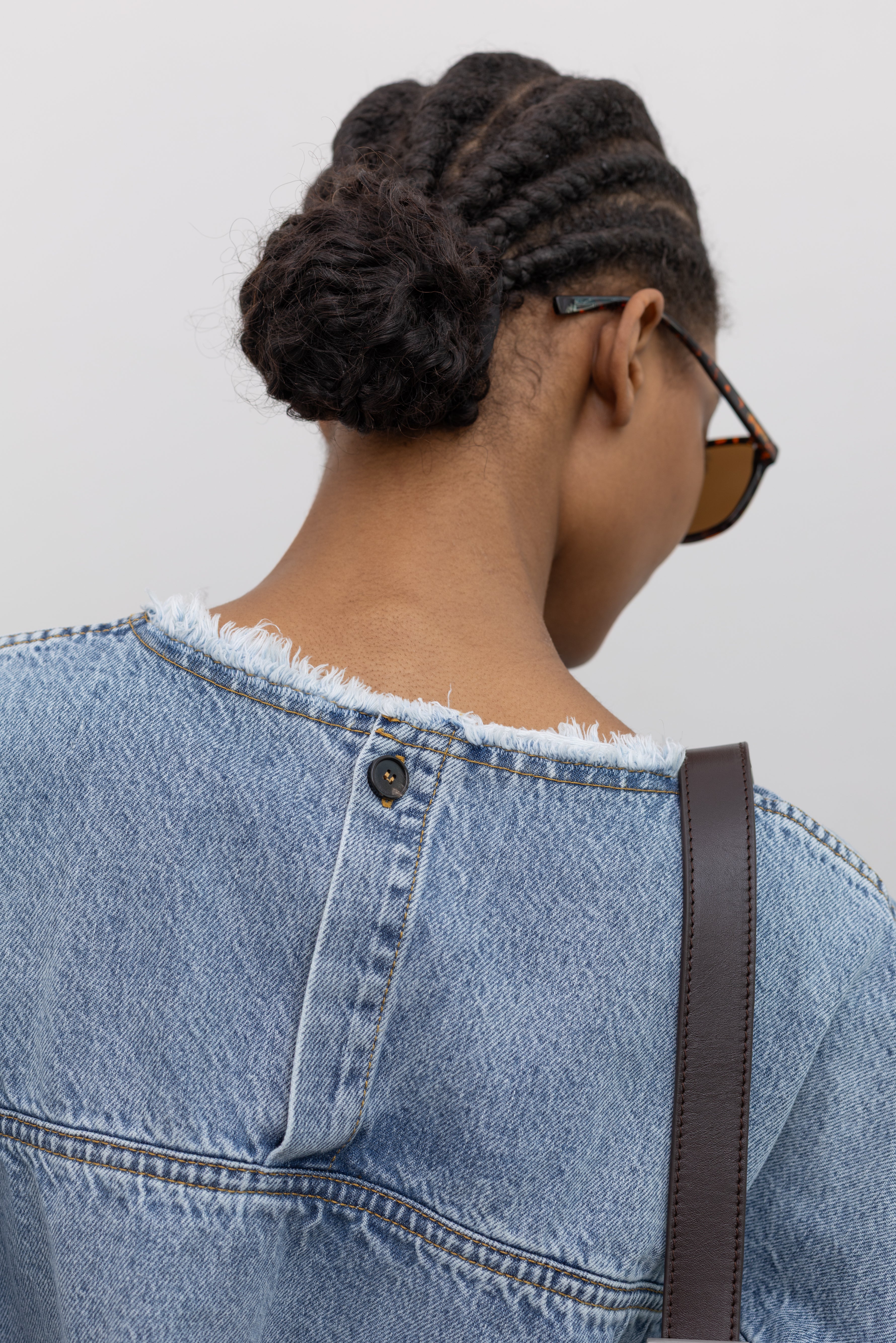 Close-up back view of a woman wearing an elevated and feminine spring denim top by Tela featuring an oversized short-sleeve, a clean light-blue wash, distinct raw-edge detailing, and a minimalist back neck closure at an Alas Eius store. 