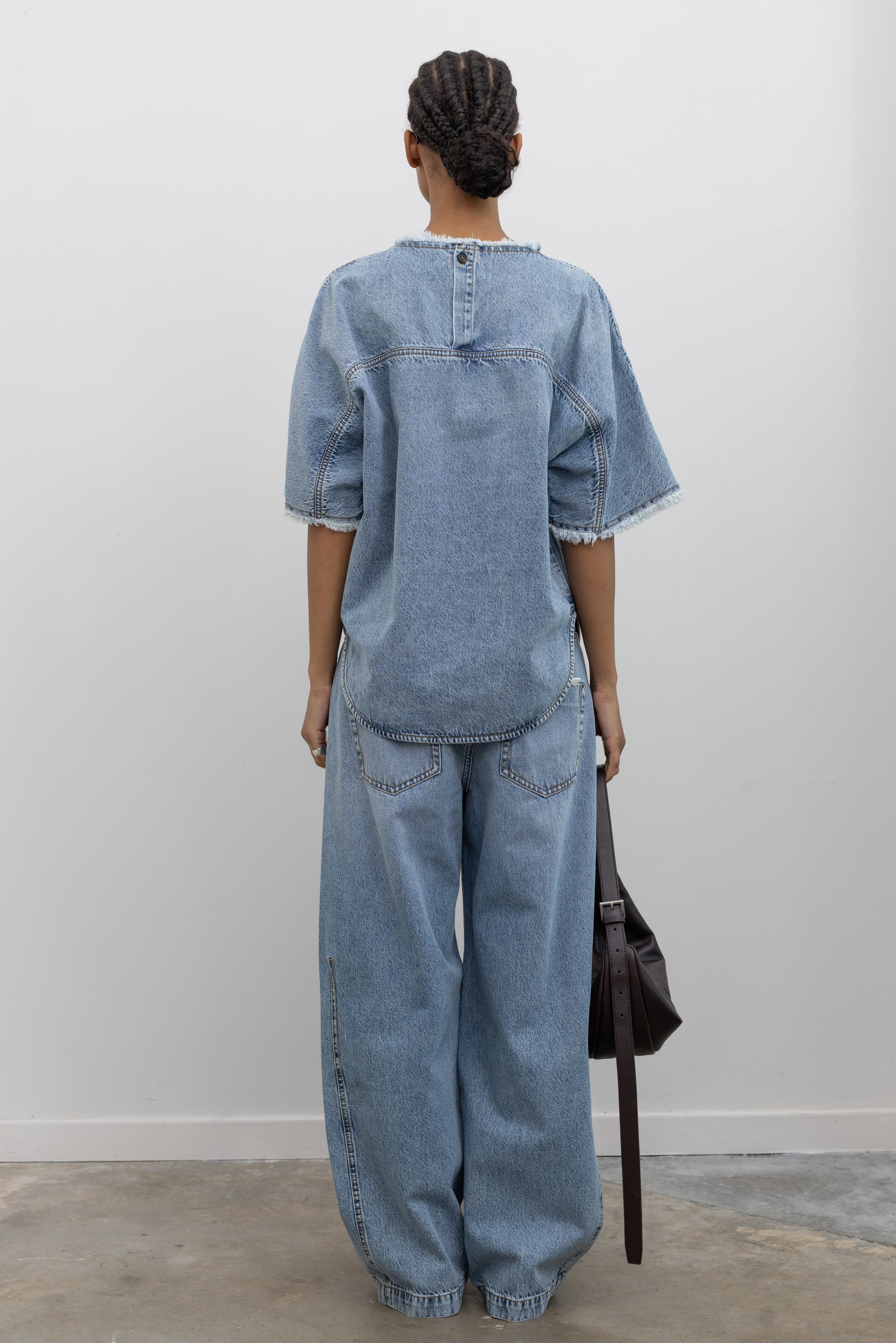 Full length back view of a Woman wearing chic spring streetwear fashion in a Tela Light Blue Denim Oversized Short Sleeve Shirt featuring  a clean light-blue wash, distinct raw-edge detailing, and a minimalist back neck closure paired with Tela light-blue wash baggy denim jeans at Alas Eius. 