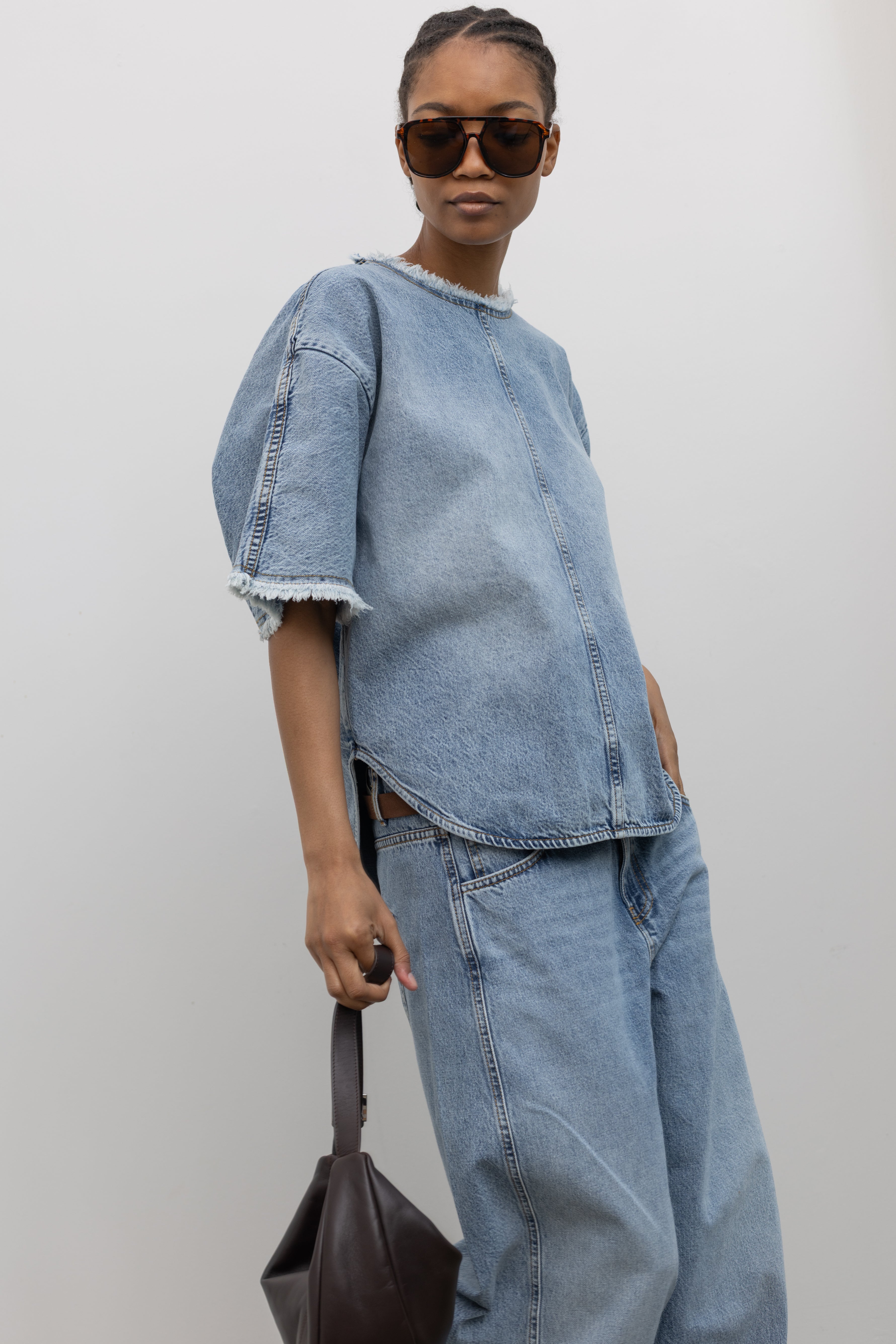3/4 view of a Woman posing with sunglasses and a St. Agni leather bag wearing an elevated and chic spring denim top by Tela featuring an oversized short-sleeve, a clean light-blue wash, distinct raw-edge detailing, and a minimalist back neck closure paired with light-blue wash denim jeans at Alas Eius. 