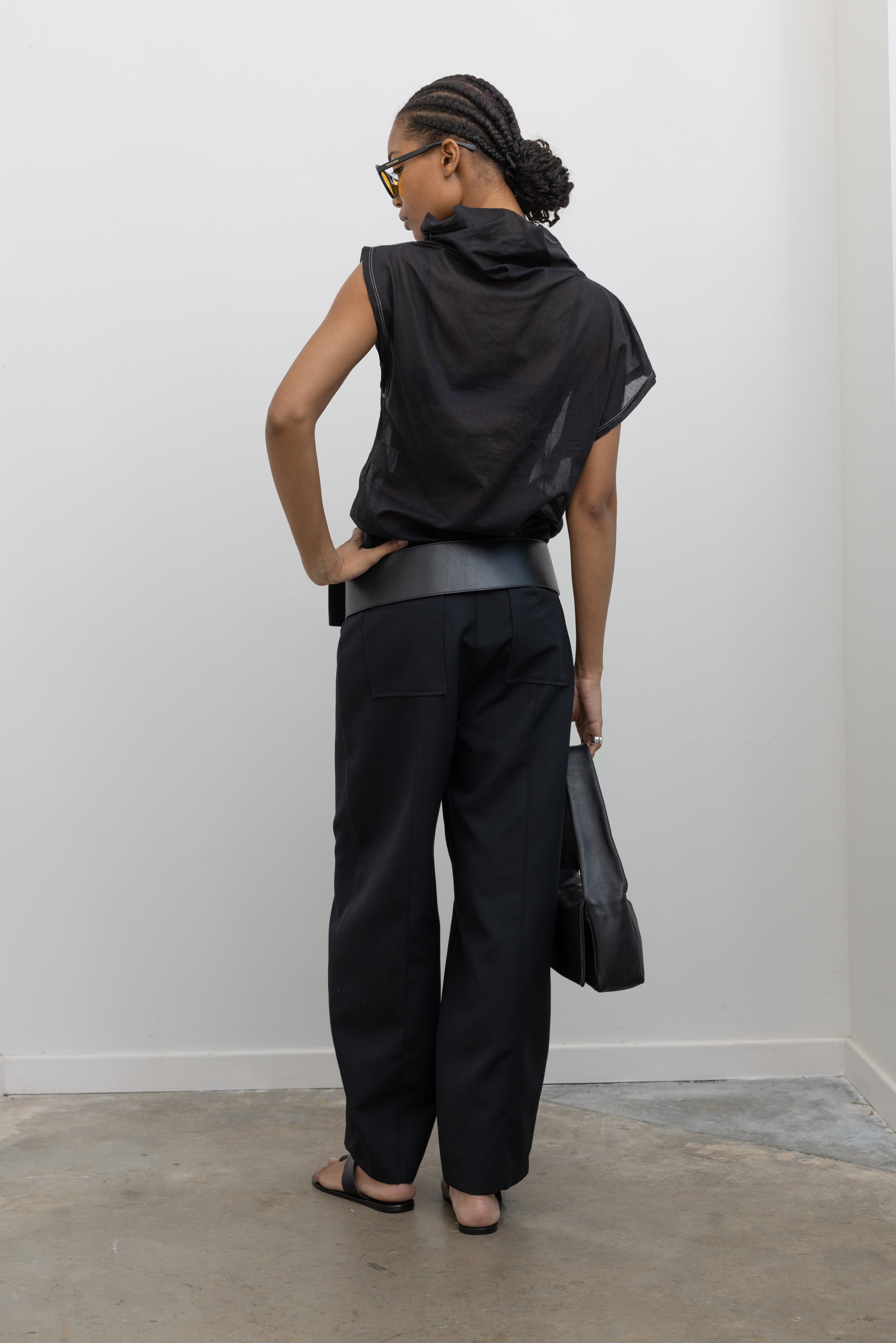 Full length back view of a woman posing wearing modern minimalist spring fashion in a Tela semi-sheer lightweight washed muslin black top featuring a soft draped asymmetrical neckline paired with a classic wide black leather belt on black wide leg trousers at an Alas Eius store. 