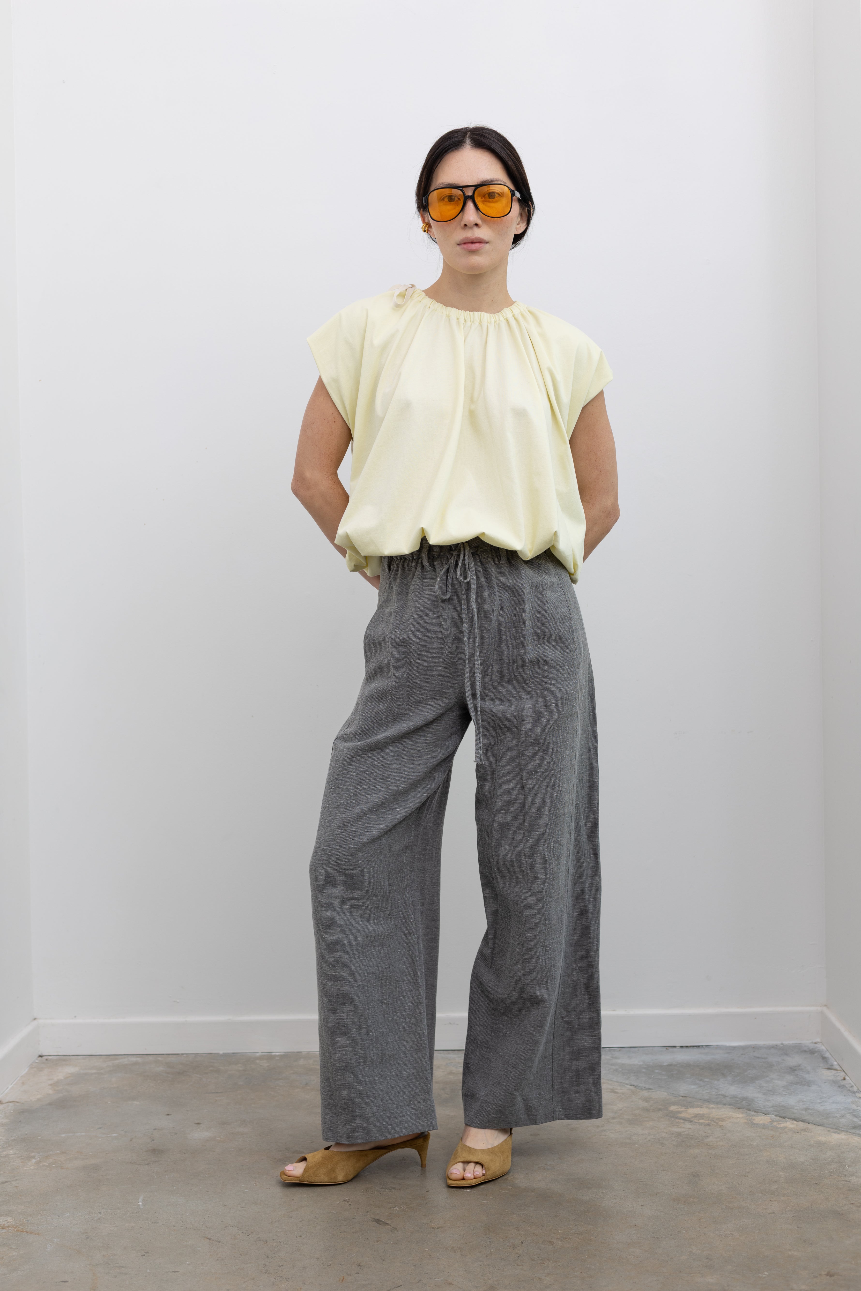 Full length front view of a woman standing wearing modern minimalist spring fashion in a Tela Light Yellow Gathered Neck Bubble Hem Top, Tela Lucio light gray drawstring pants featuring a wide leg and side seam pockets, and Tela Feng Mustard Leather Heeled Sandals at an Alas Eius store. 