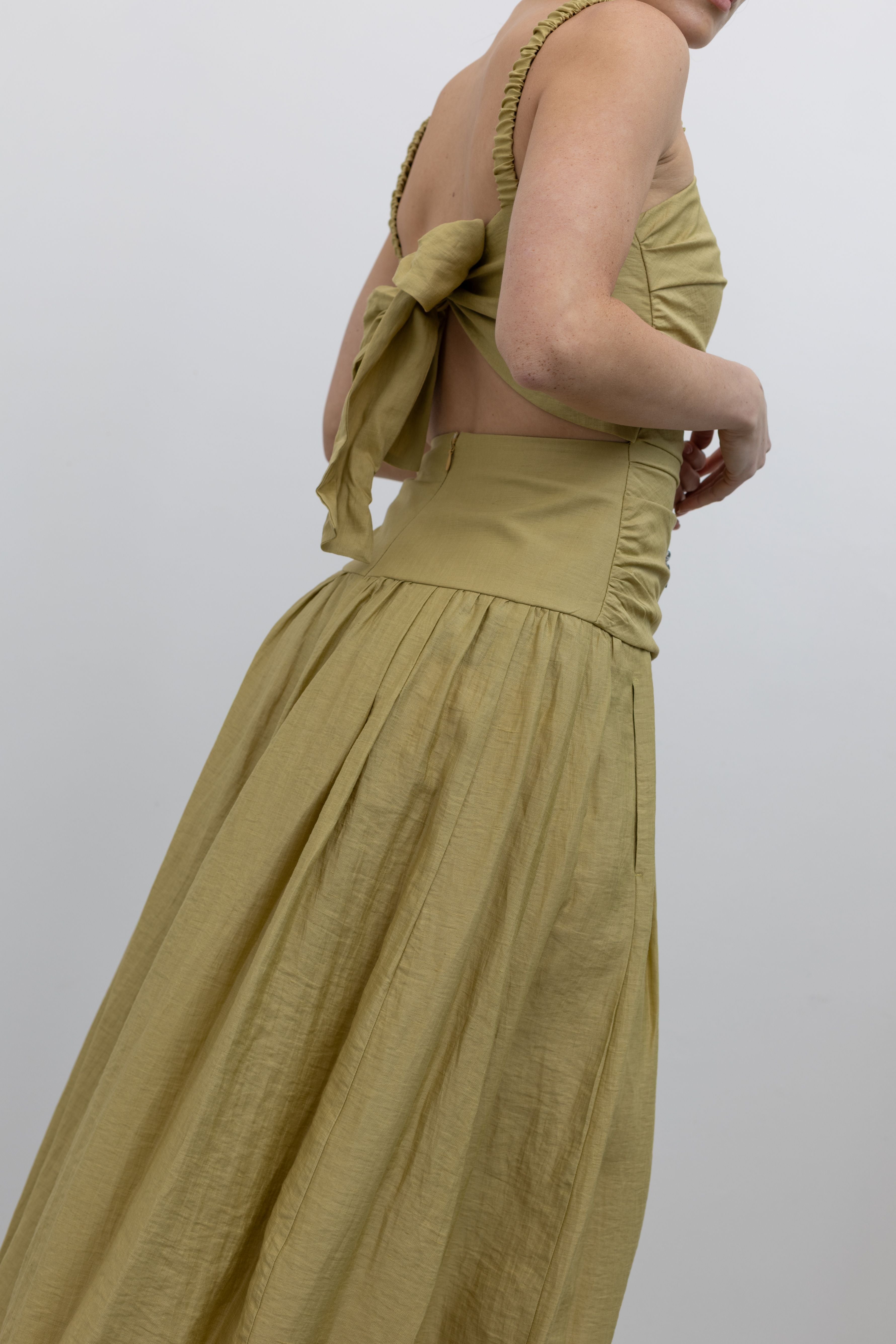 Close-up side view of a woman standing wearing chic and feminine spring fashion in a Tela label linen blend pistachio green maxi dress with flattering ruching, delicate narrow shoulder straps, and a striking back cut-out detail with a bow closure paired with white leather slingback kitten heels at an Alas Eius store. 