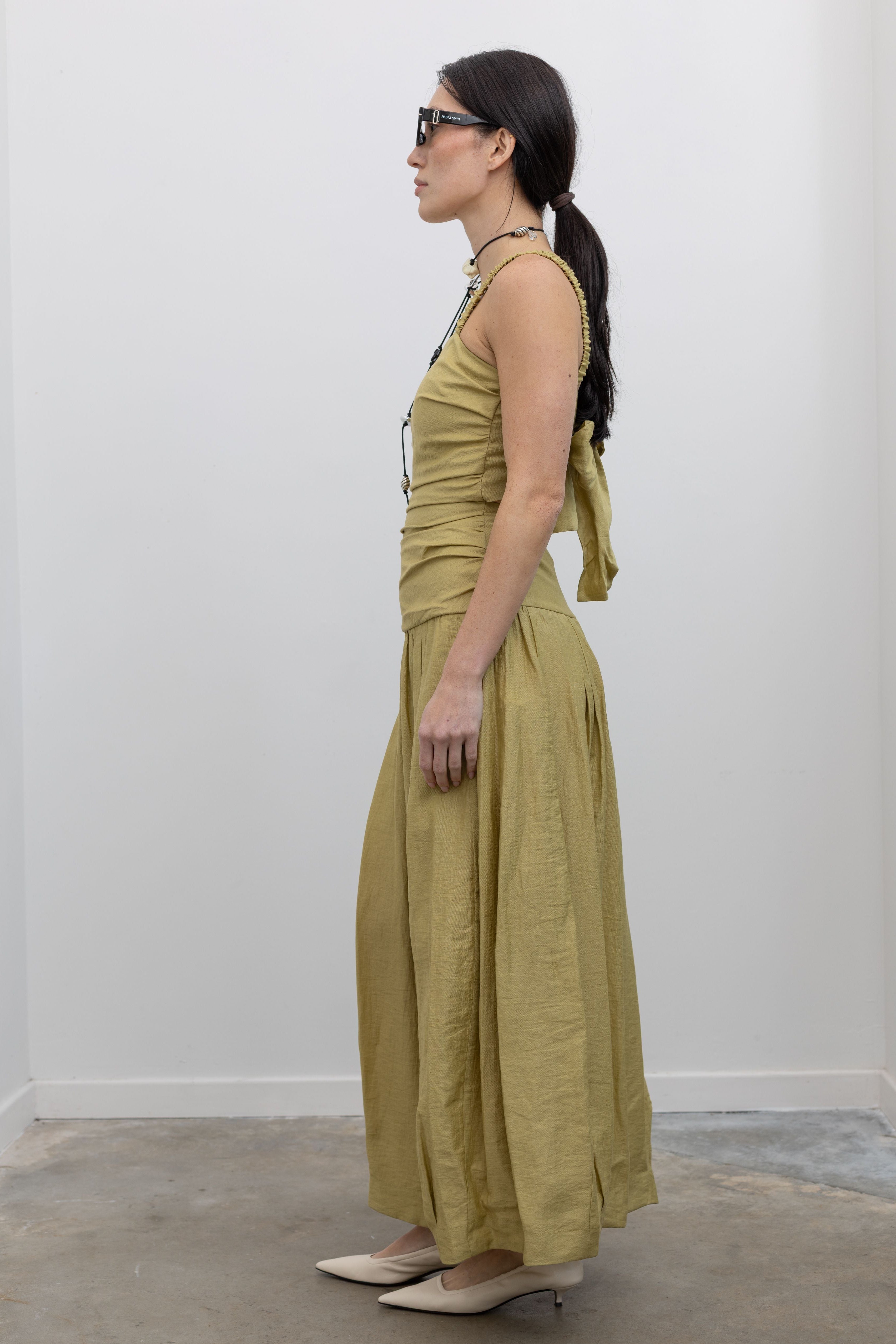 Full length side view of a woman standing wearing modern and elegant spring fashion in a Tela label linen blend pistachio green maxi dress with flattering ruching, delicate narrow shoulder straps, and a striking back cut-out detail with a bow closure paired with white leather slingback kitten heels at an Alas Eius store. 