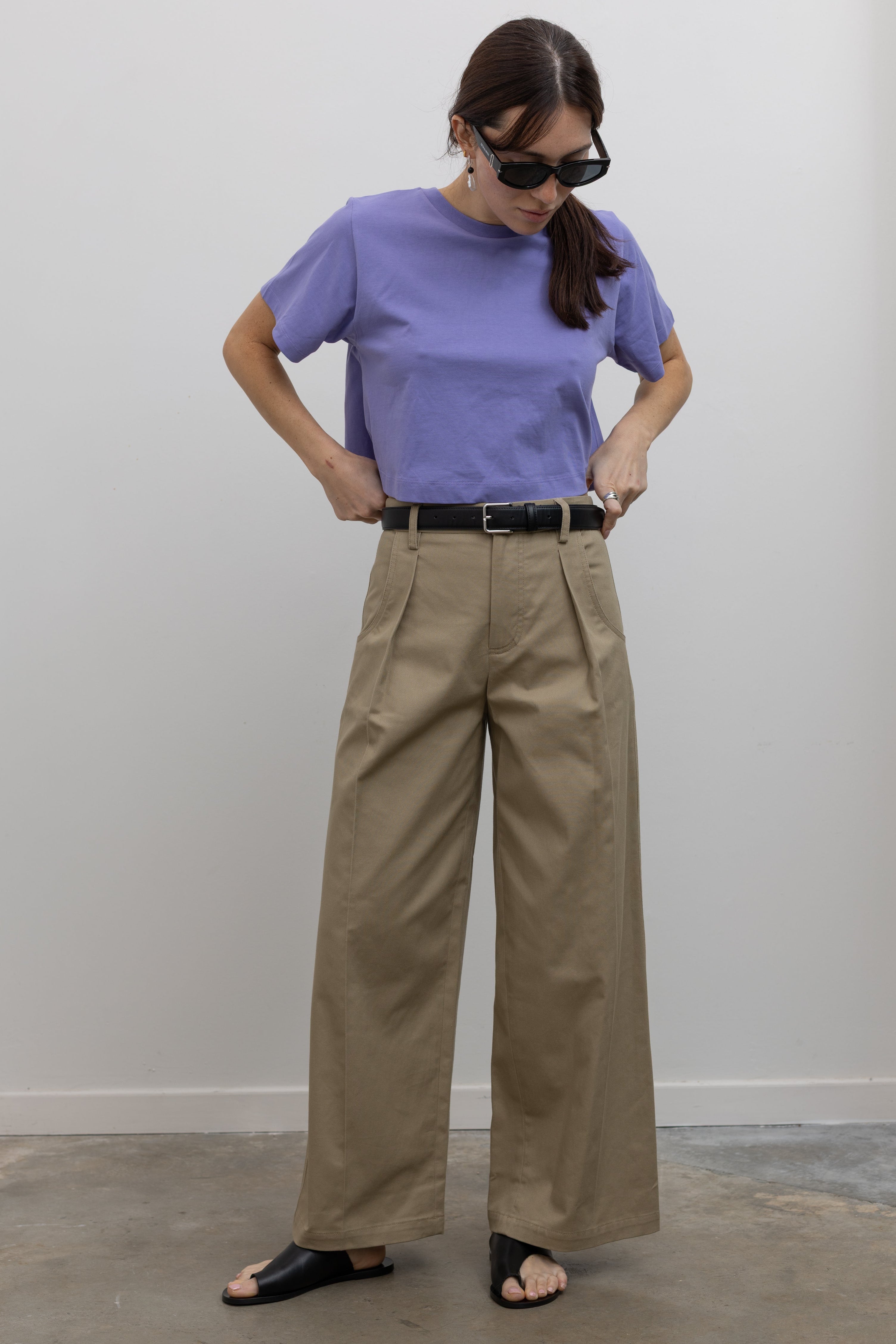 Woman standing wearing Spring fashion in a pair of Tela Rubicon Sand Cotton Gabardine Trousers and a blue tee with sunglasses and sandals at Alas Eius